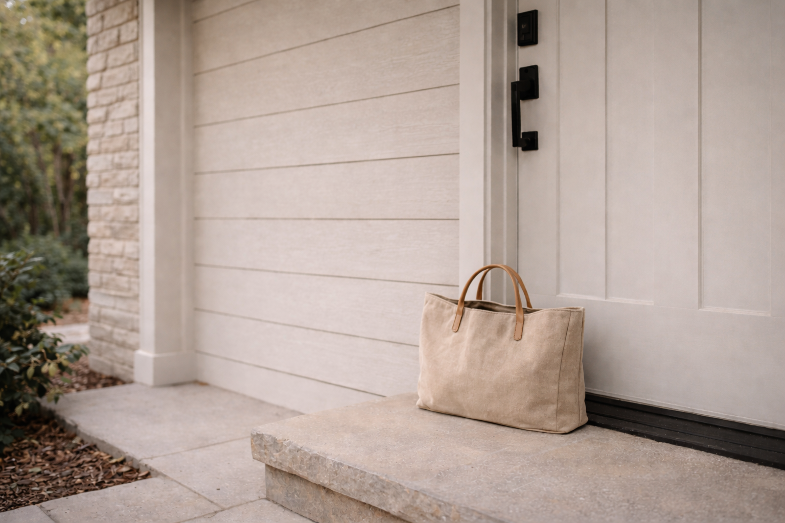 A beige tote bag with leather straps resting on a concrete step outside a house.