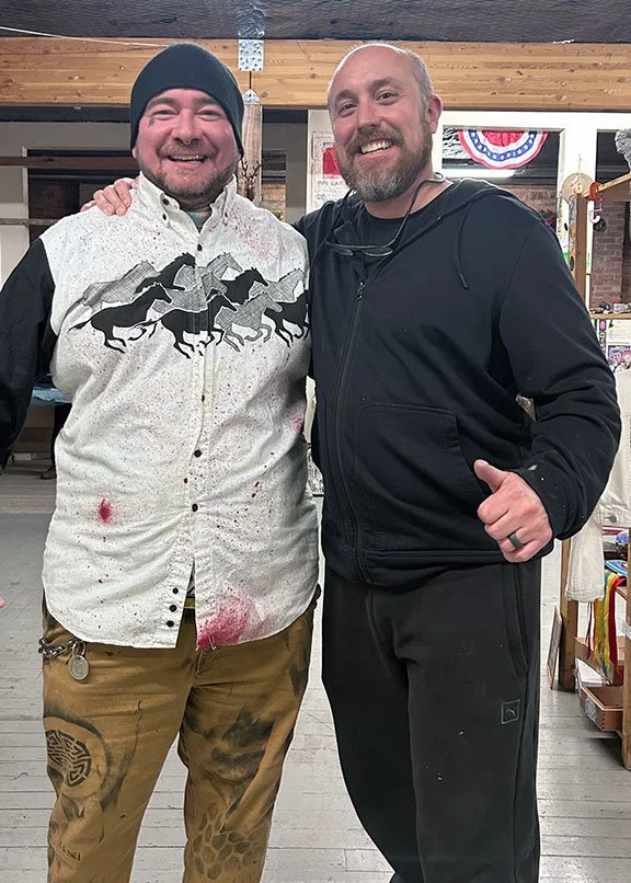 Two smiling men posing indoors, one wearing a black beanie and a white shirt with horse graphics, the other in a black jacket with glasses around his neck. They have their arms around each other.