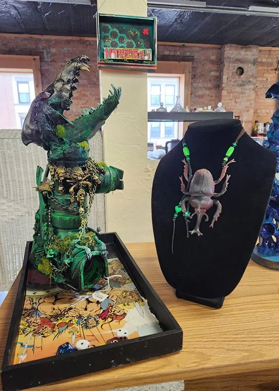 A toy dinosaur figure dressed in green, adorned with chains and jewelry, holding a small object, and placed on a tray with dice and game pieces. Next to it, a mounted model of a spider decorated with green beads is displayed on a black stand.