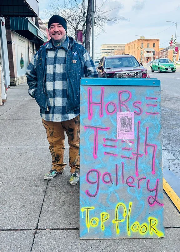 A man standing on a sidewalk next to a colorful sign that reads 'Horse Teth gallery Top floor' with a flyer attached, with cars and buildings in the background.