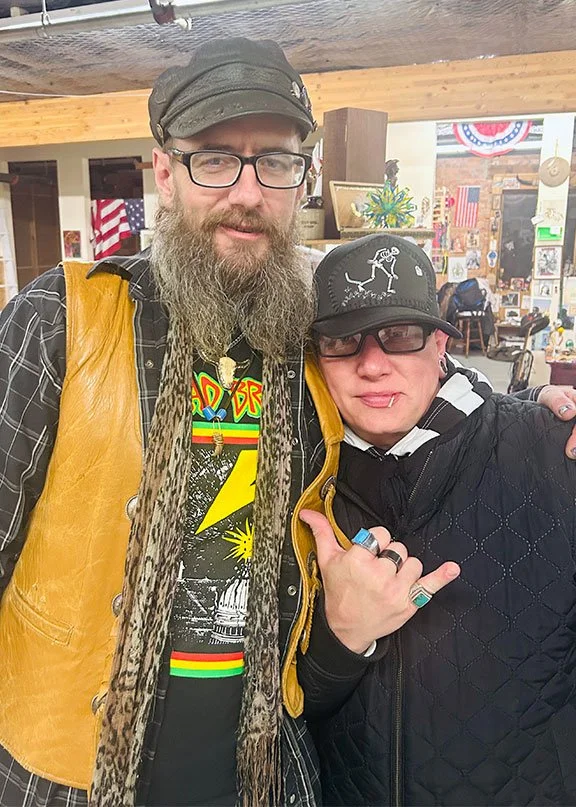 Two men posing together in a decorated indoor space, one with a beard wearing glasses, a black cap, a yellow vest, and a graphic t-shirt. The other with shorter hair wearing glasses, a black cap with white embroidery, and a black quilted jacket making a hand gesture.