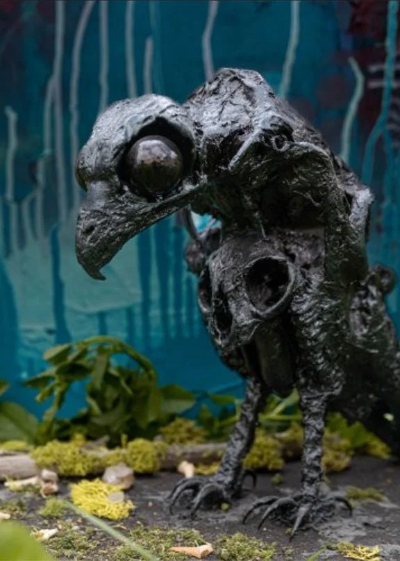 A sculpture of a bird with a large beak and round eyes, set outdoors with moss, plants, and a blue background.
