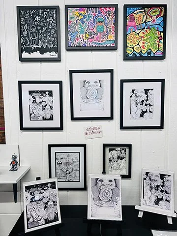 Gallery of framed artwork on a white wall, featuring colorful and black-and-white illustrations, with a small table and additional art pieces on stands in front.