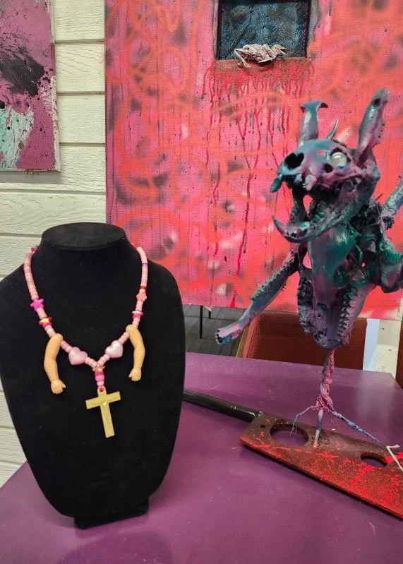 A black jewelry display bust with a pink beaded necklace that has a yellow cross pendant hanging from it. Next to the bust is a creature sculpture with a dog skull head, multiple eyes, and a body resembling a bird or rat, standing on a red-painted wooden base. In the background, there is an abstract pink and orange painting with drips and a small window.