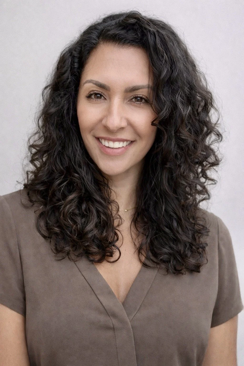 A woman with long, curly dark hair smiling, wearing a brown V-neck top and a delicate necklace, against a plain light background.