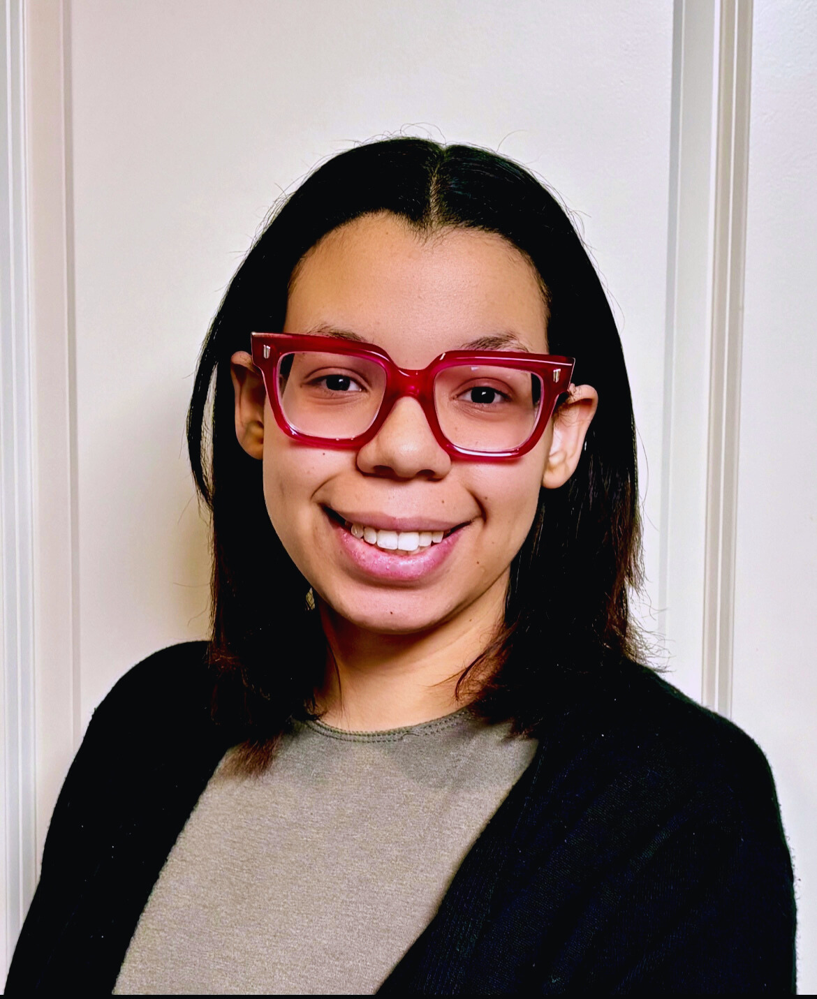 A young woman with shoulder-length dark hair, wearing large red glasses, smiling, standing in front of a white wall.