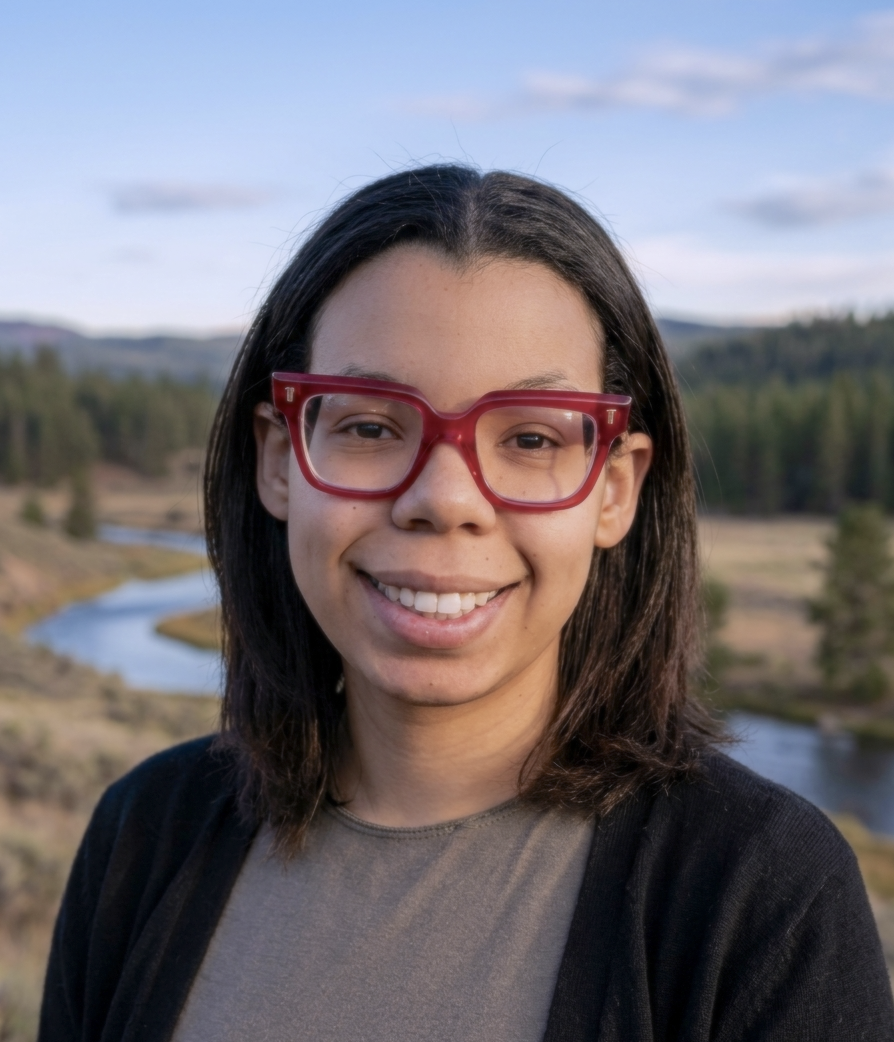 A young woman with shoulder-length dark hair, wearing large red glasses, smiling, standing in front of a white wall.