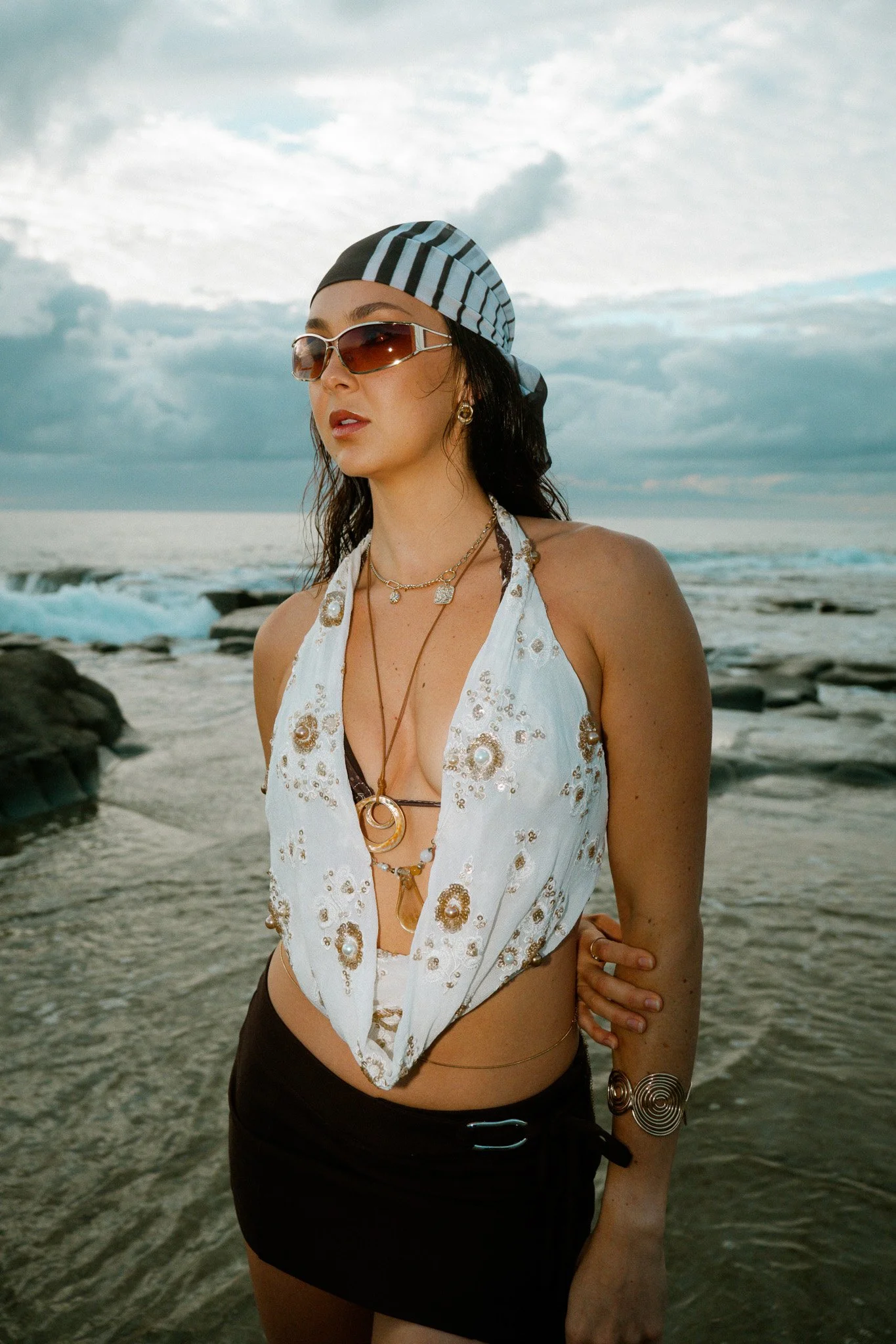 Woman wearing sunglasses, bandana, and jewelry on a beach with rocks and waves.
