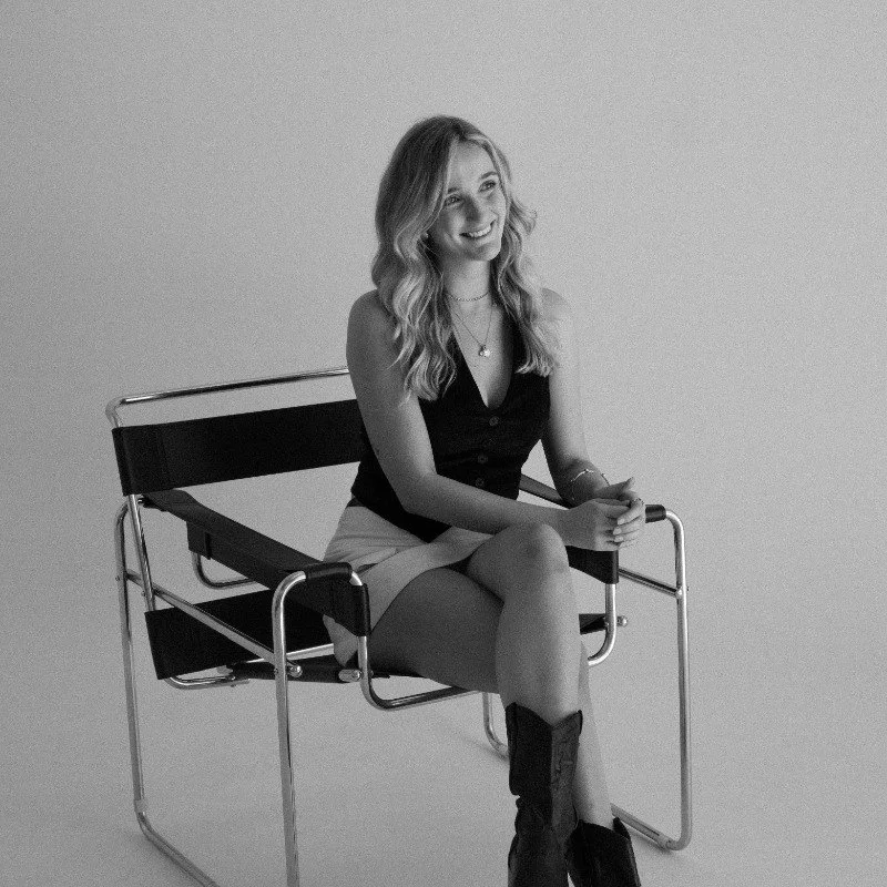 Black and white photo of a smiling woman sitting on a modern chair, wearing a sleeveless top, shorts, and boots, with long wavy hair and layered necklaces, against a plain background.