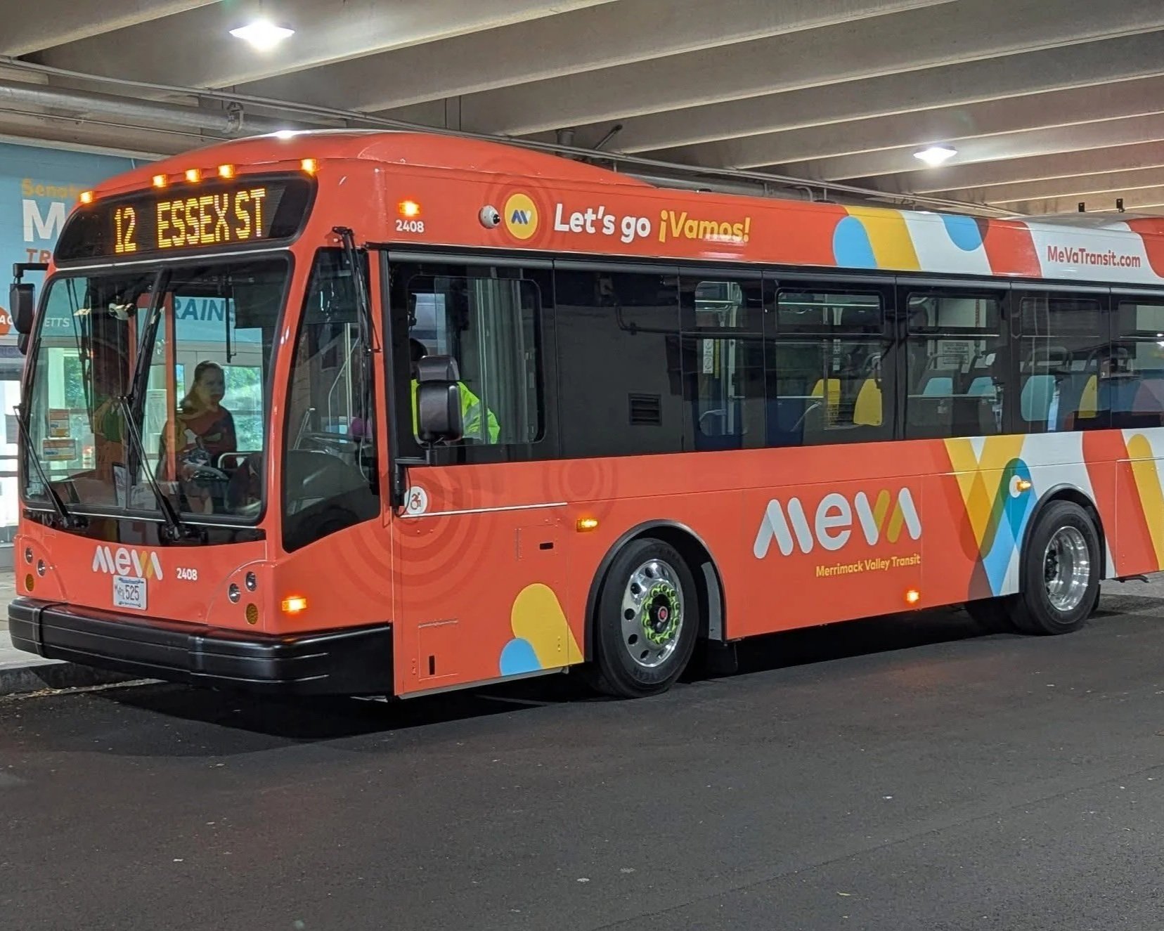 A colorful Merrimack Valley Transit bus parked underground, with digital sign displaying route 12 to Essex St, and branding including the words "Let's go ¡Vamos!" and the logo of ALeXA.