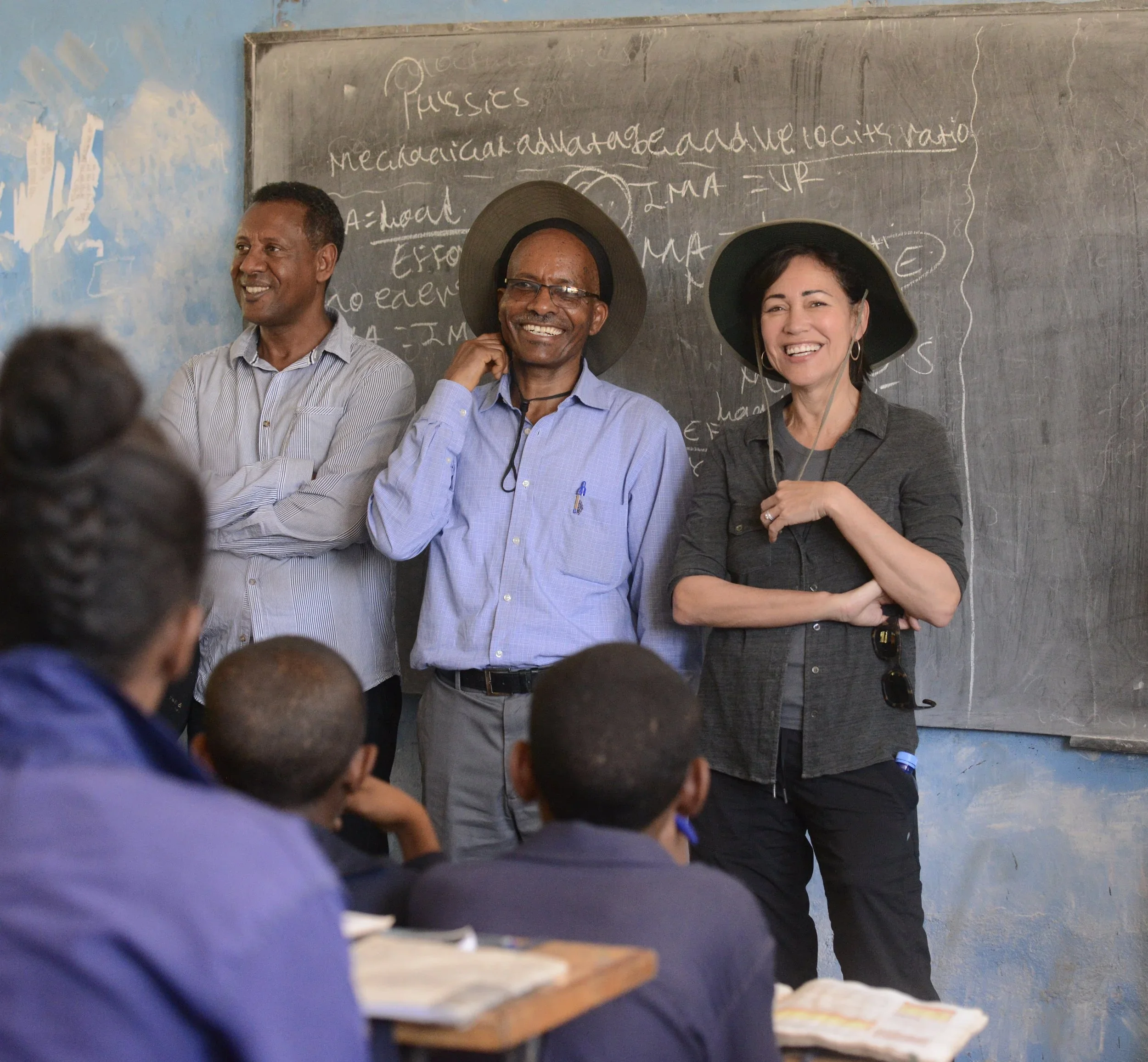 Three teachers standing in front of a classroom with students seated at desks, smiling and wearing hats, in a rural school classroom.