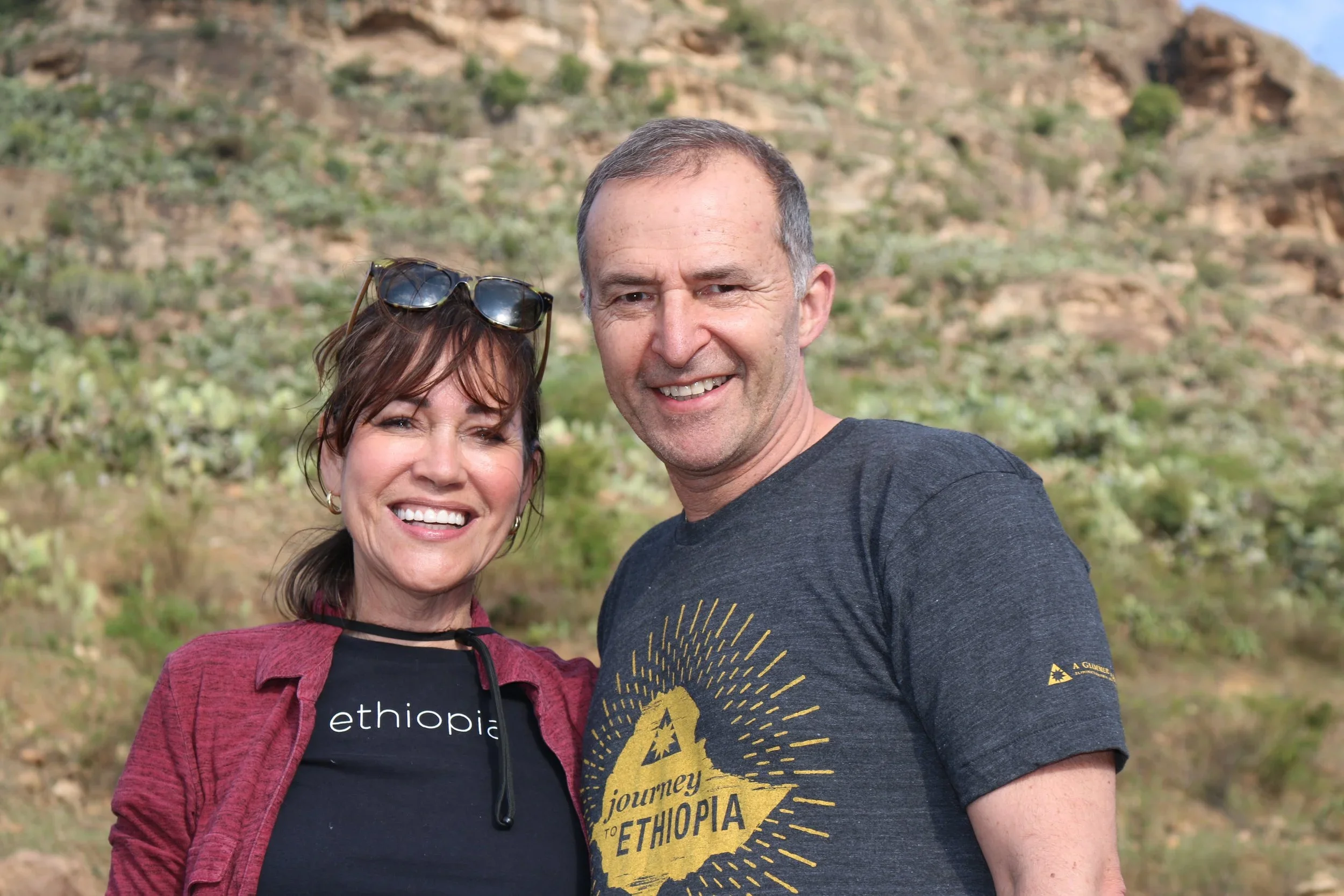 A smiling couple stands outdoors in front of a desert landscape with rocks and sparse vegetation. The woman has brown hair, sunglasses on her head, and is wearing a black t-shirt and a red jacket. The man has short gray hair, and is wearing a dark gr