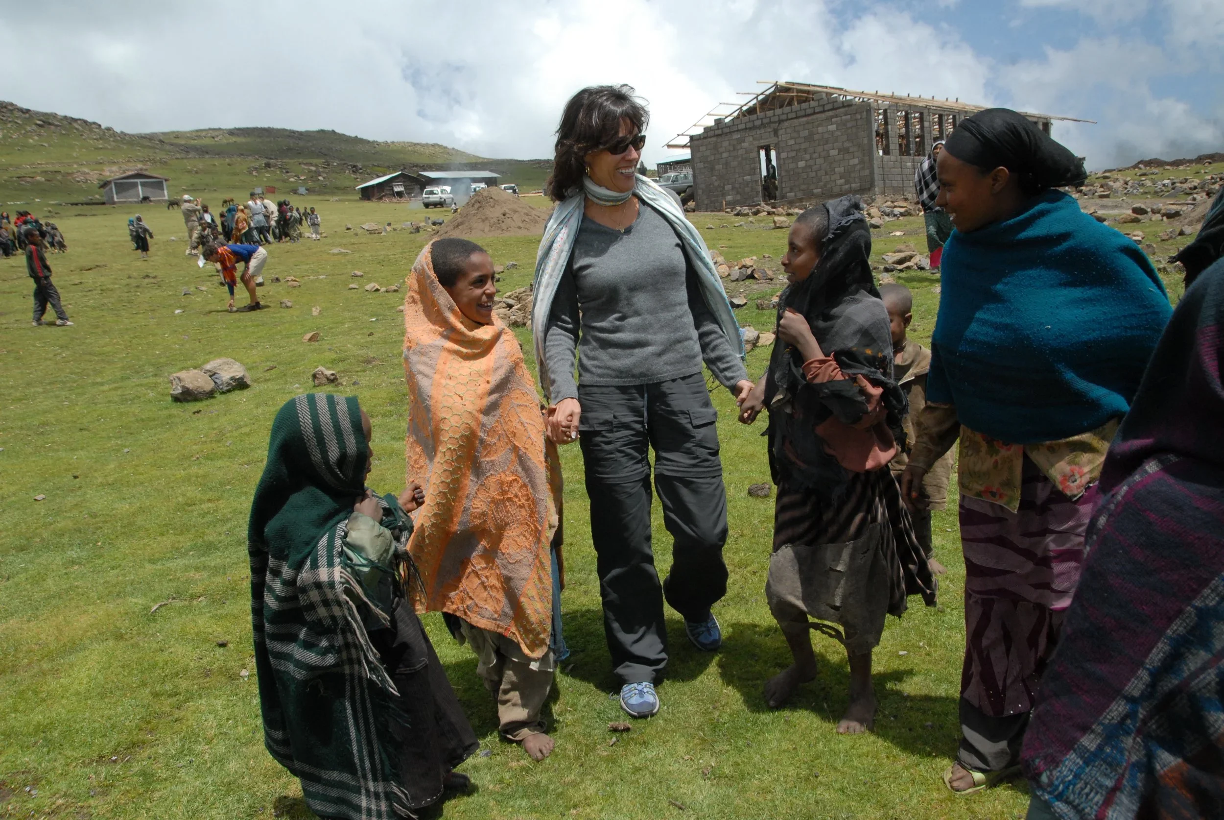 A woman holding hands with children in a grassy field, with a construction site and other people in the background.