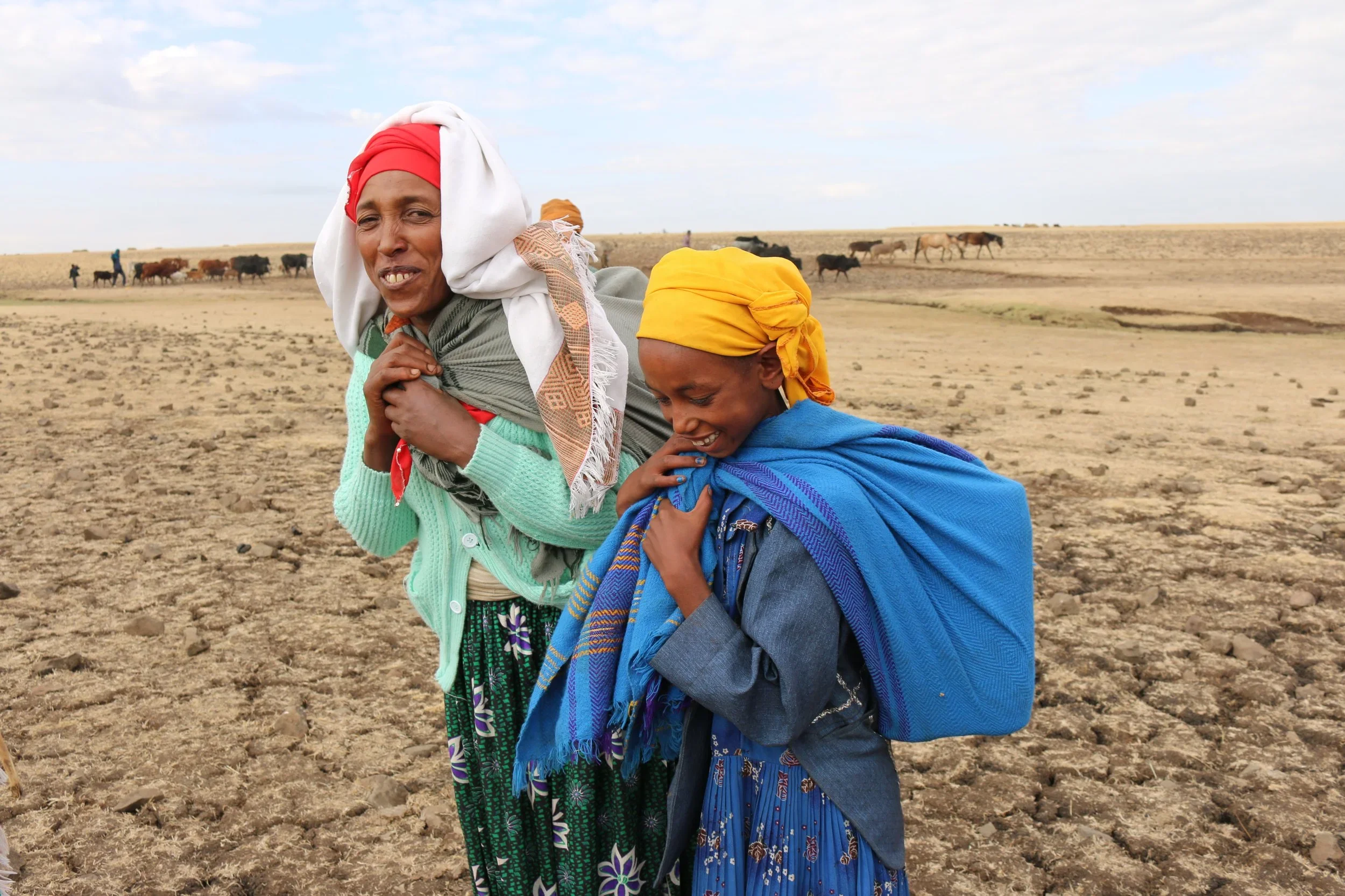 Two women smiling and standing in a dry, cracked field, with cattle in the background, wearing colorful headscarves and clothing.
