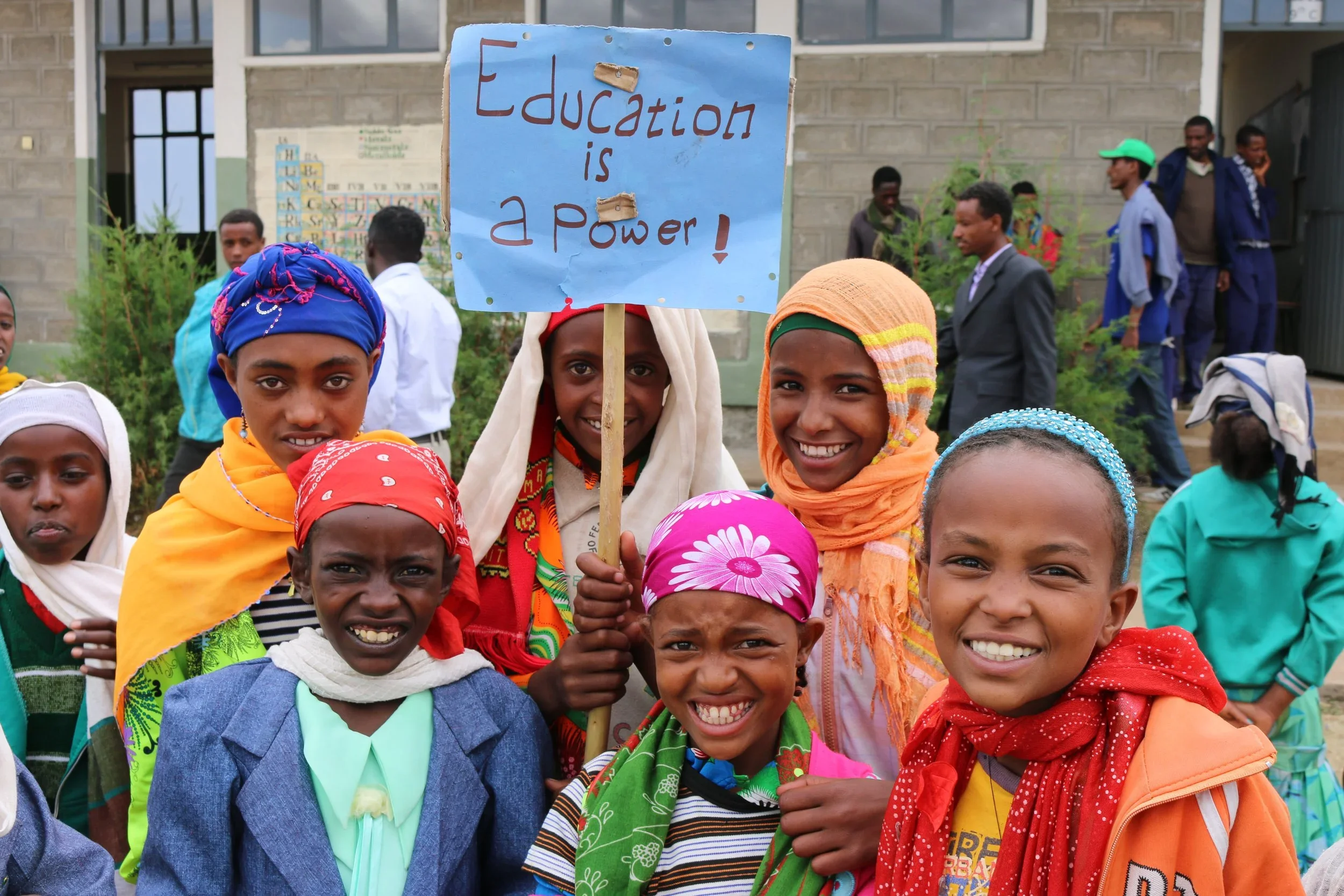 Group of smiling children holding a sign that says "Education is a Power!" in front of a school building.