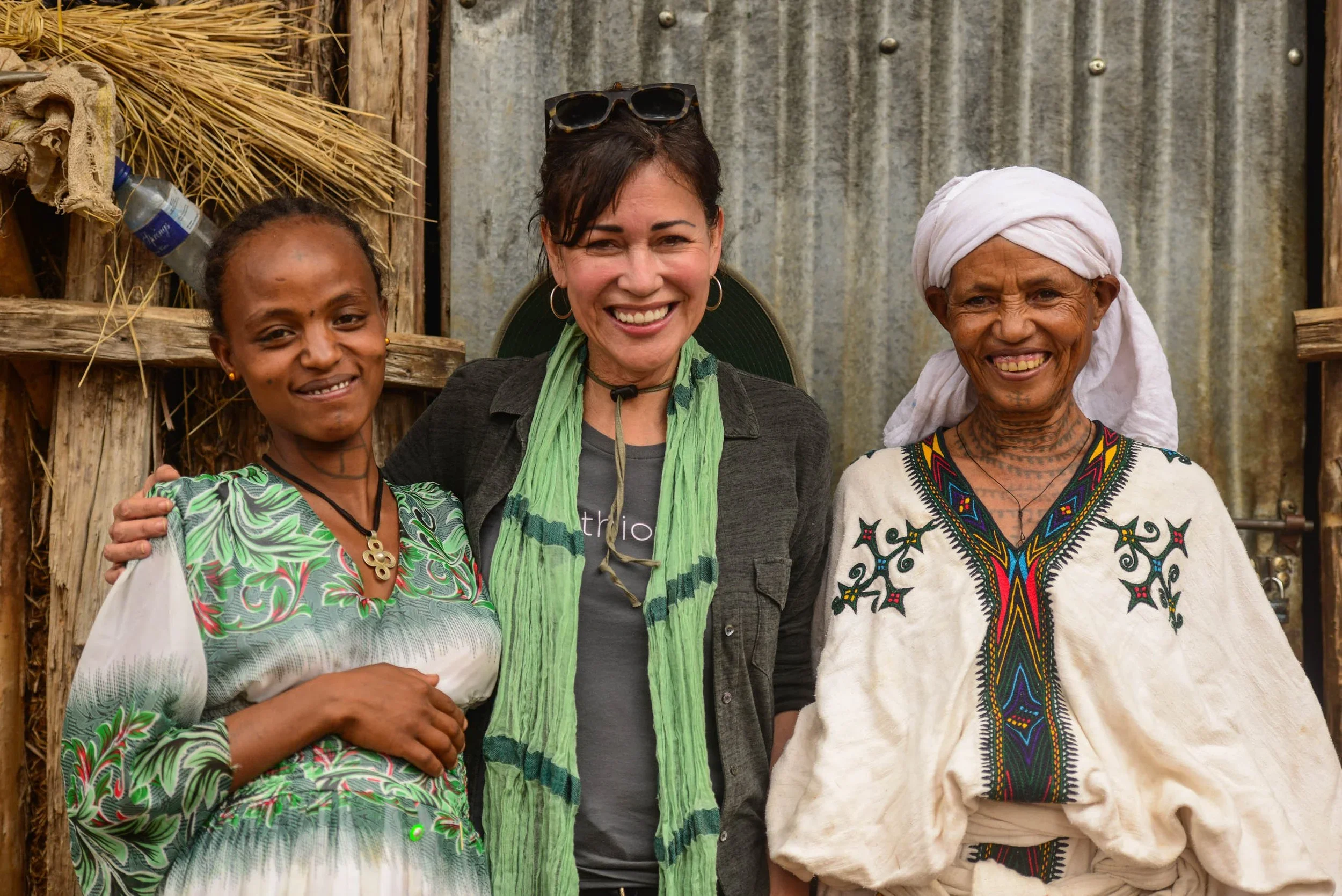 Three women standing close together, smiling, outside with a rustic background of wood and metal.