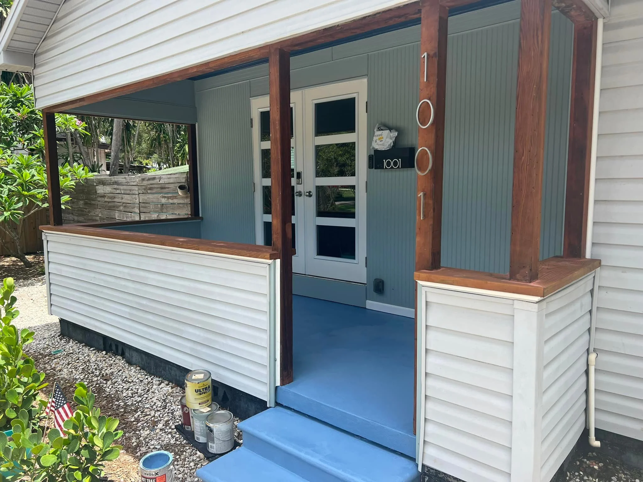 Front porch of a house with white siding, a small blue staircase leading to the front door, and wooden posts framing the porch. There are paint cans on the ground near the stairs, and a mailbox labeled "1001" next to the door.