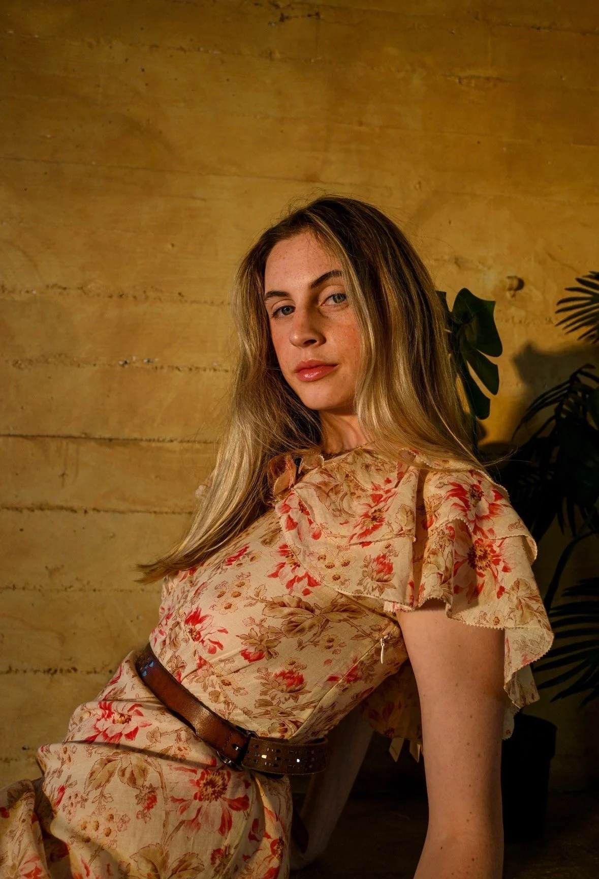A woman with long blonde hair wearing a floral dress with ruffled sleeves, sitting against a yellow wooden wall with green leafy plants in the background. model, stellina, stella spound, sustainable fashion, second hand clothing, designer vintage rar