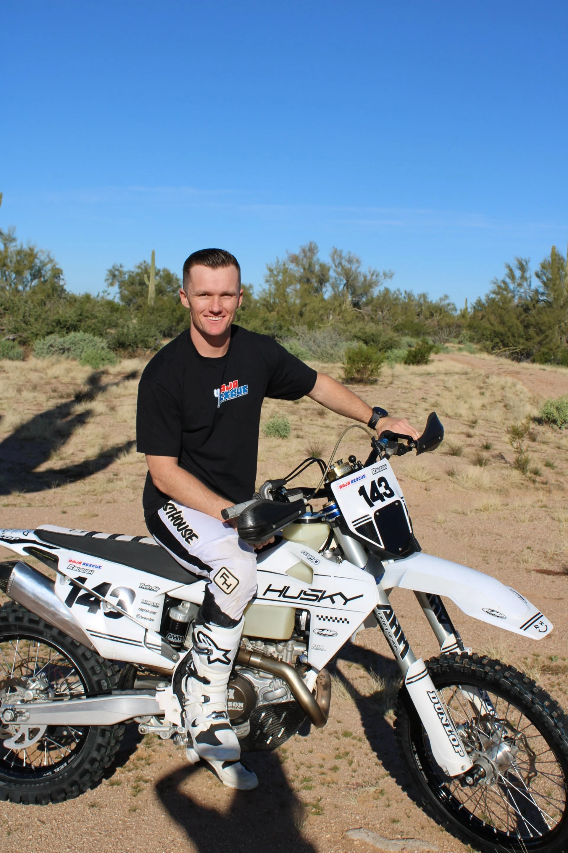 A young man smiling while sitting on a white Husqvarna dirt bike in a desert landscape with sparse vegetation and a clear blue sky.