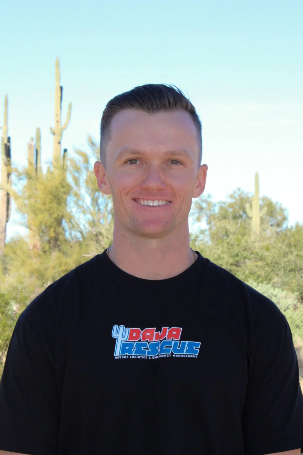 A young man in a police uniform holding a framed certificate, smiling, with a window and greenery in the background.