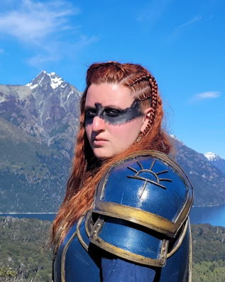 Mujer con maquillaje oscuro en la cara, armadura y peinado con trenzas, en un paisaje montañoso con lago y picos nevados.