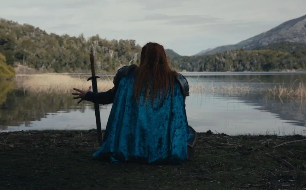 Persona con capa azul sentado junto a un lago rodeado de árboles y montañas.