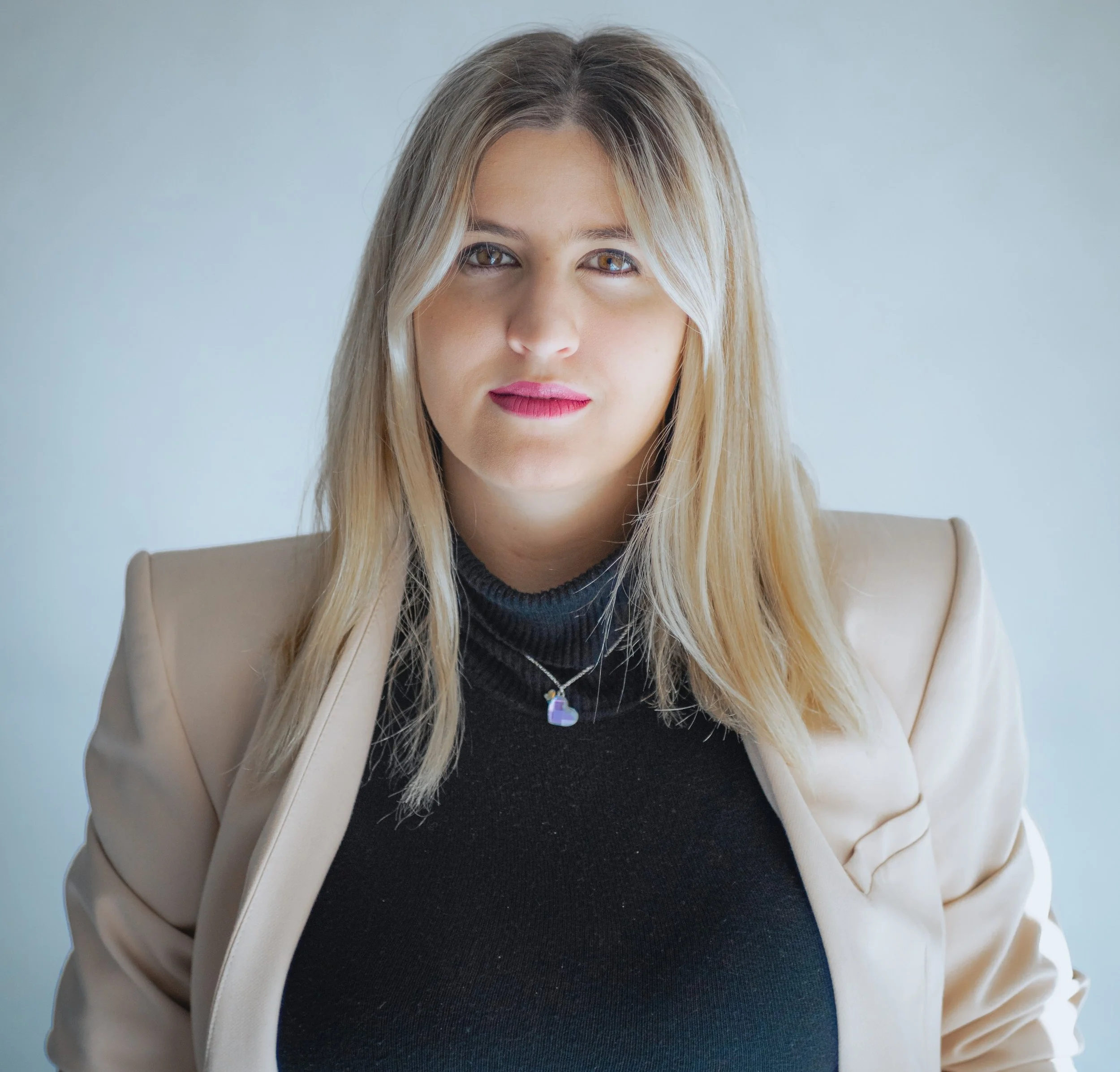 Retrato de una mujer de cabello rubio y ojos color miel, usando un blazer crema, blusa negra y collar de corazón, frente a un fondo gris claro.
