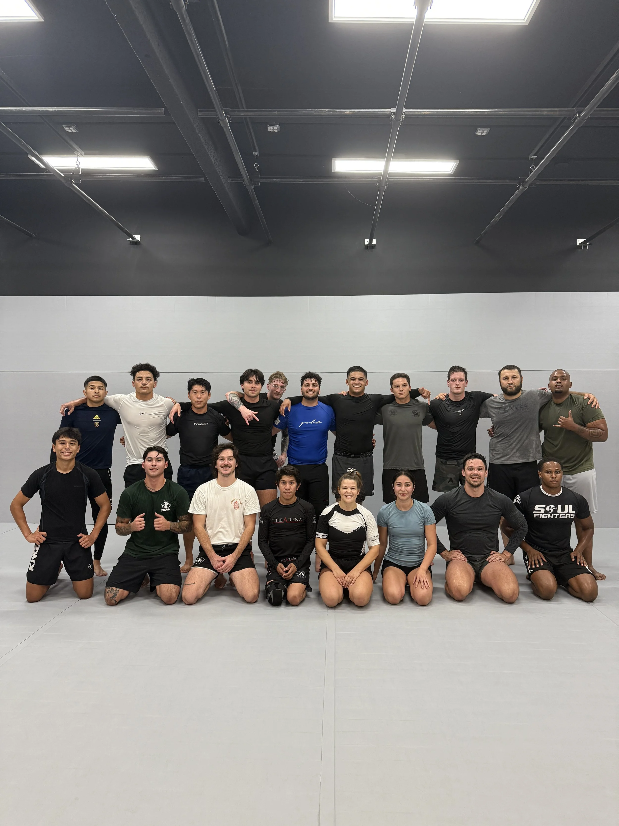 Group of martial artists in a gym, posing for a photo, some kneeling and some standing, wearing athletic gear.