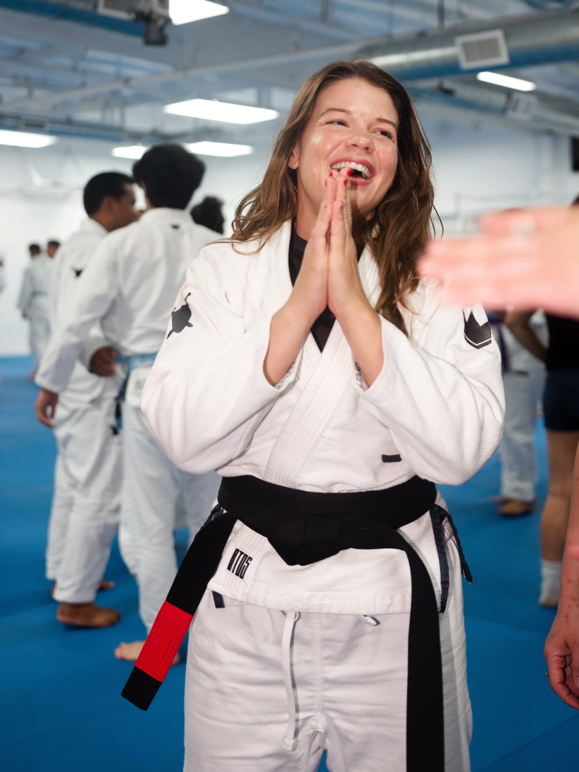A woman in a white judo gi with a black belt, smiling and holding her hands together in a praying gesture at a judo training session or competition, with other practitioners in gis in the background.