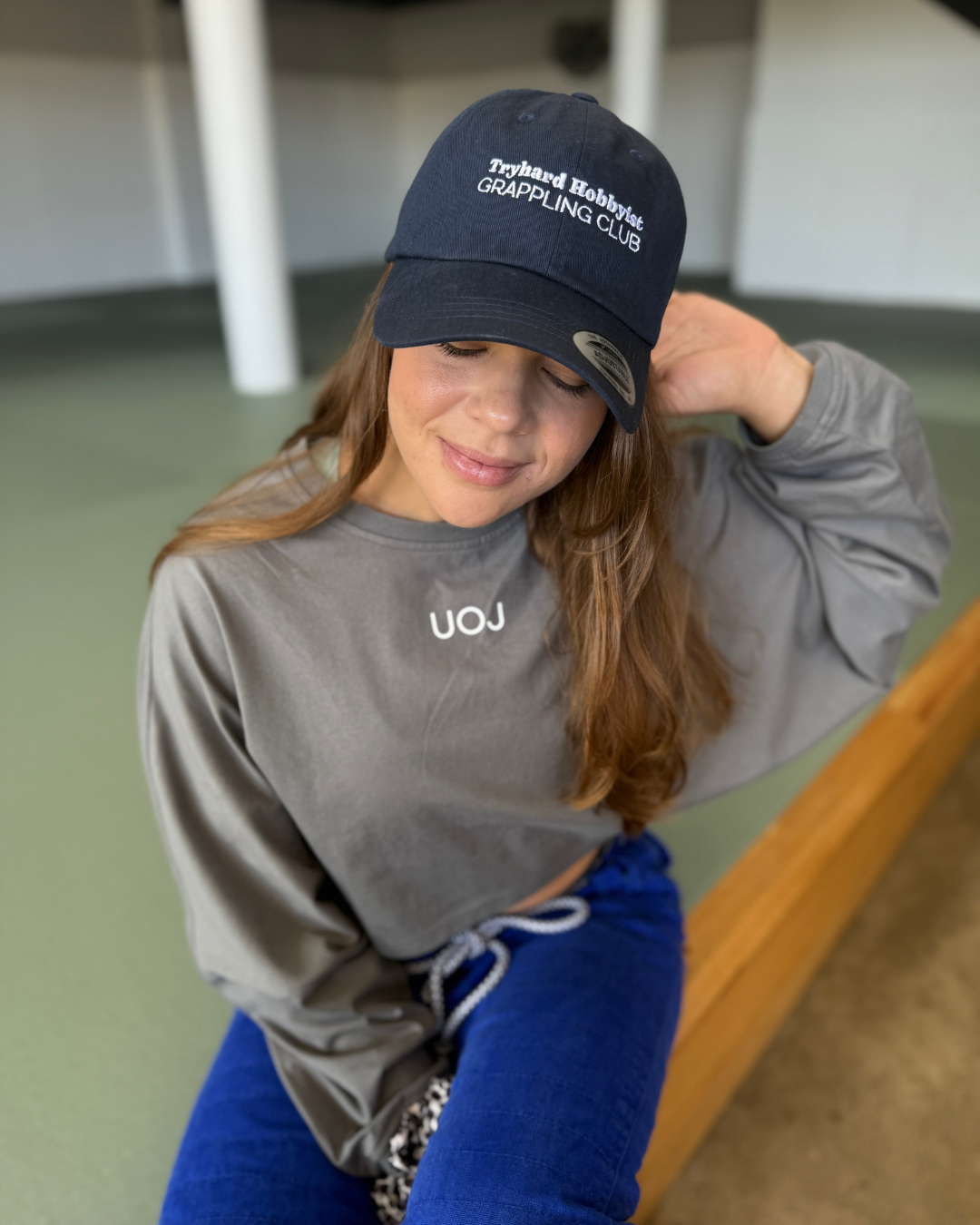 A young woman with long hair, wearing a navy baseball cap with white text, a grey sweatshirt, and blue pants, sitting on a wooden bench with her eyes closed and a gentle smile.