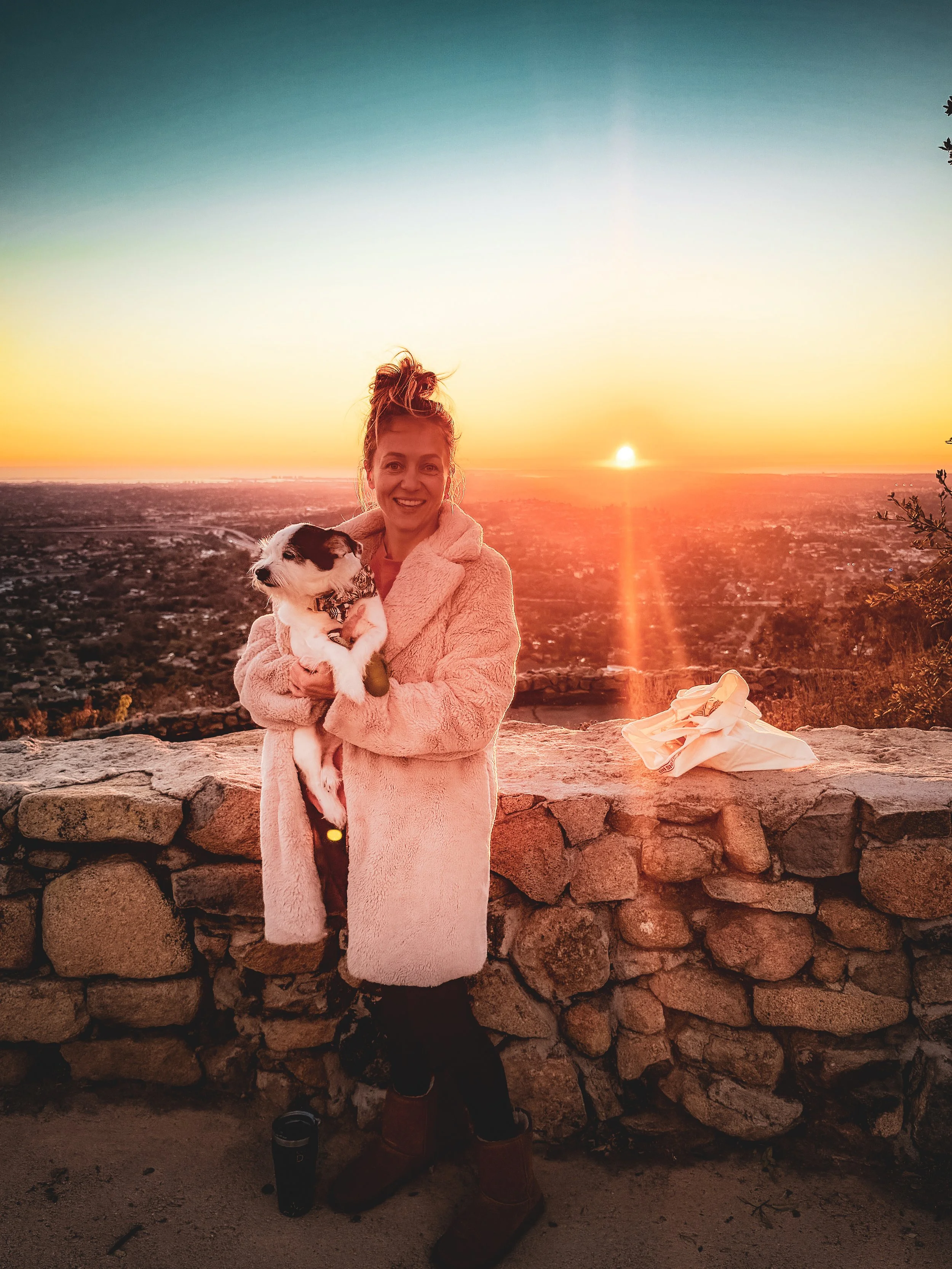 The poet, T.L. Amber, holding her dog named Lou Dog, while standing in front of a stone wall at sunset, with a sunset view over a city or landscape.