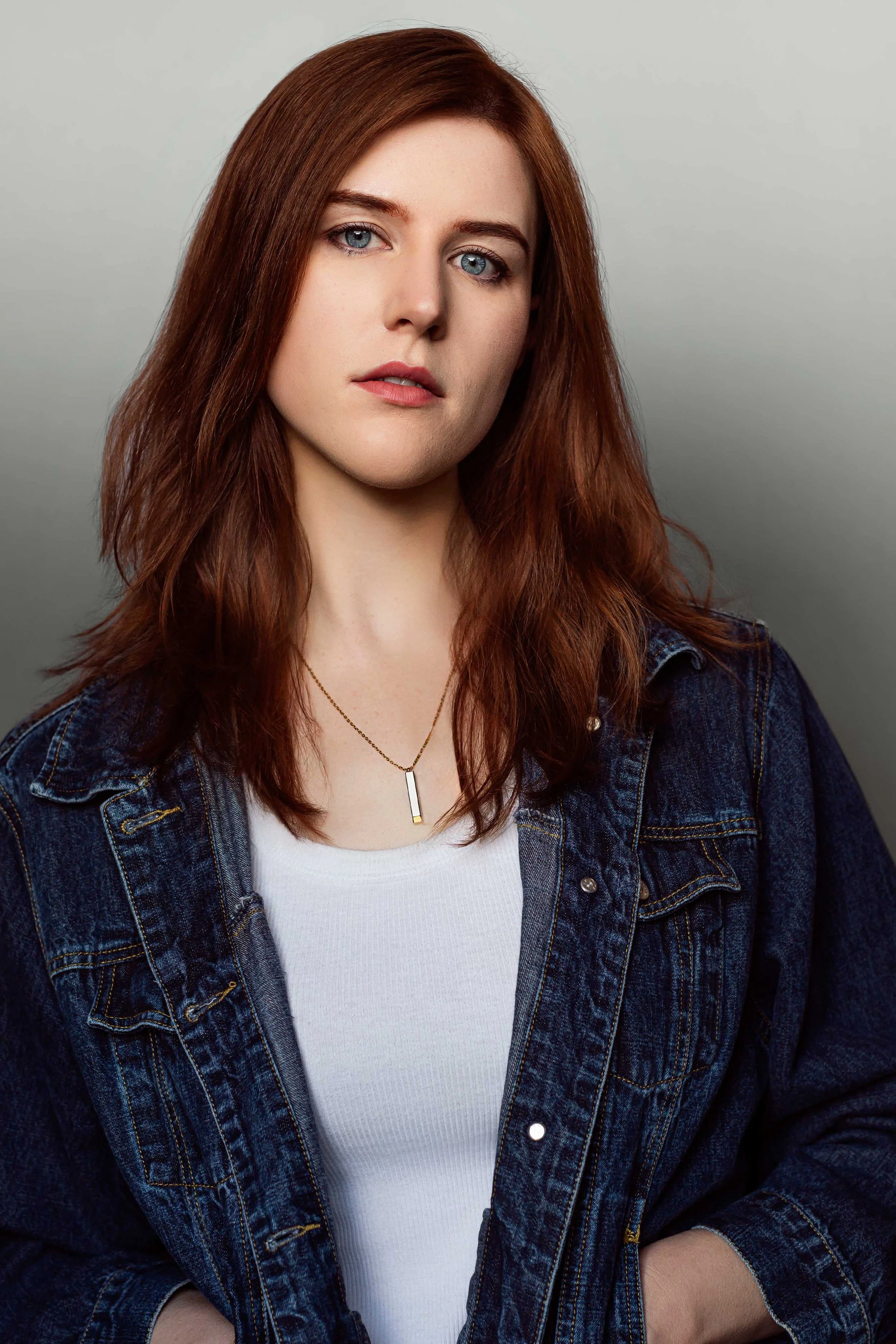 Saige Holst, a young woman with long red hair, blue eyes, wearing a white shirt, denim jacket, and a gold necklace, standing against a simple gray background.