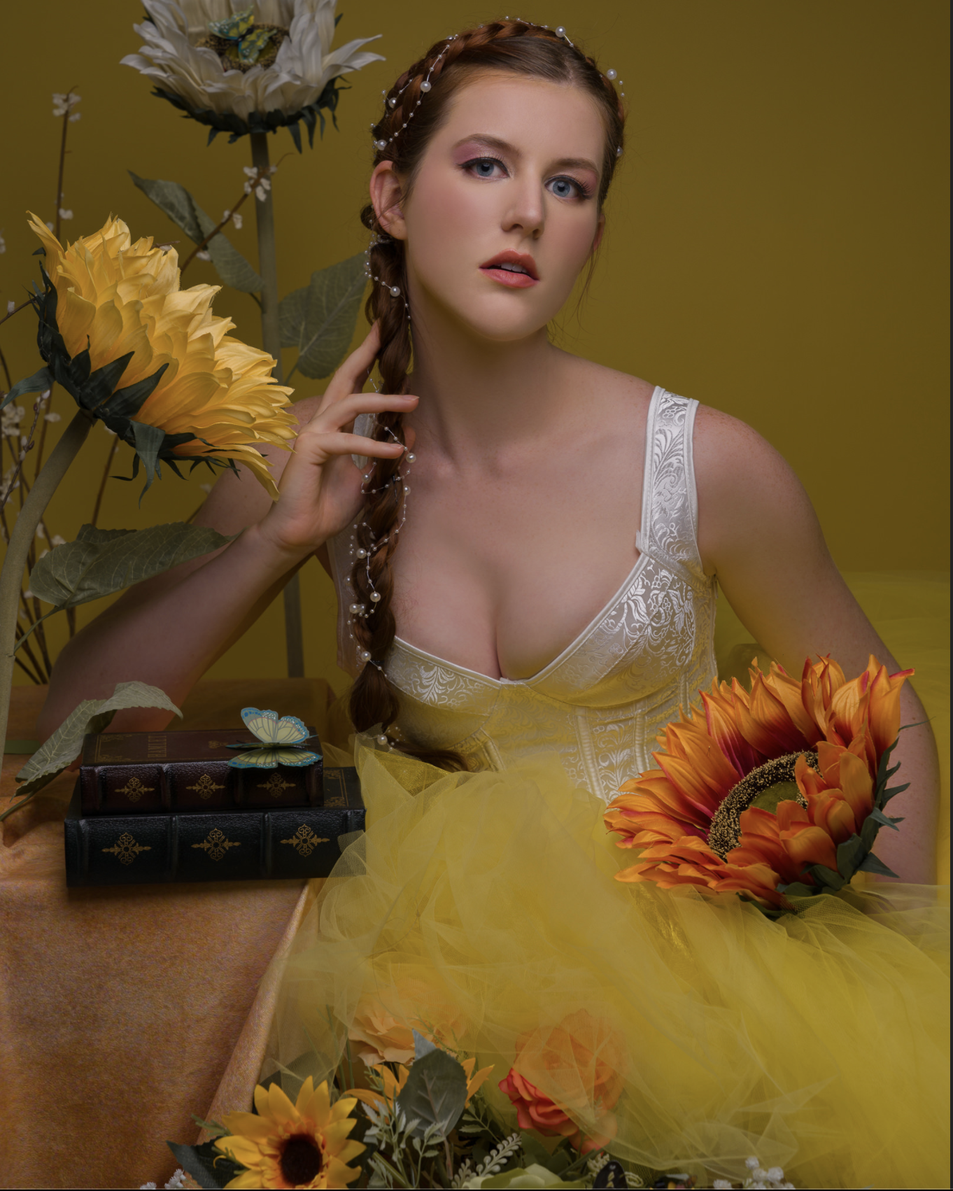 A young woman with fair skin and light blue eyes, wearing a white dress with a floral pattern, sits at a table surrounded by sunflowers and other bright flowers. She has long, red braided hair decorated with pearl strings, and a small braid on the si