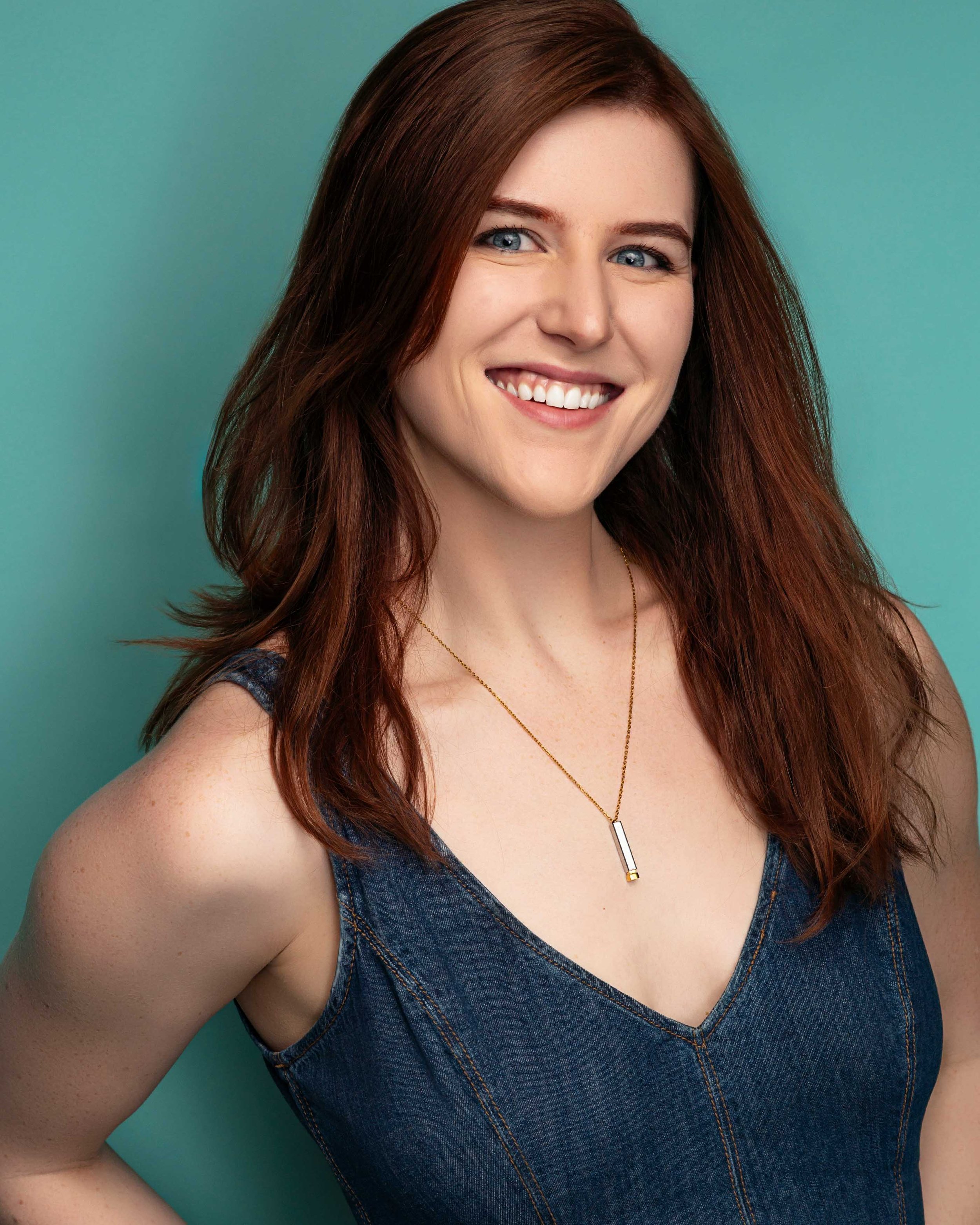Saige smiling, with long red hair, blue eyes, wearing a denim top and a gold necklace in front of a teal background.