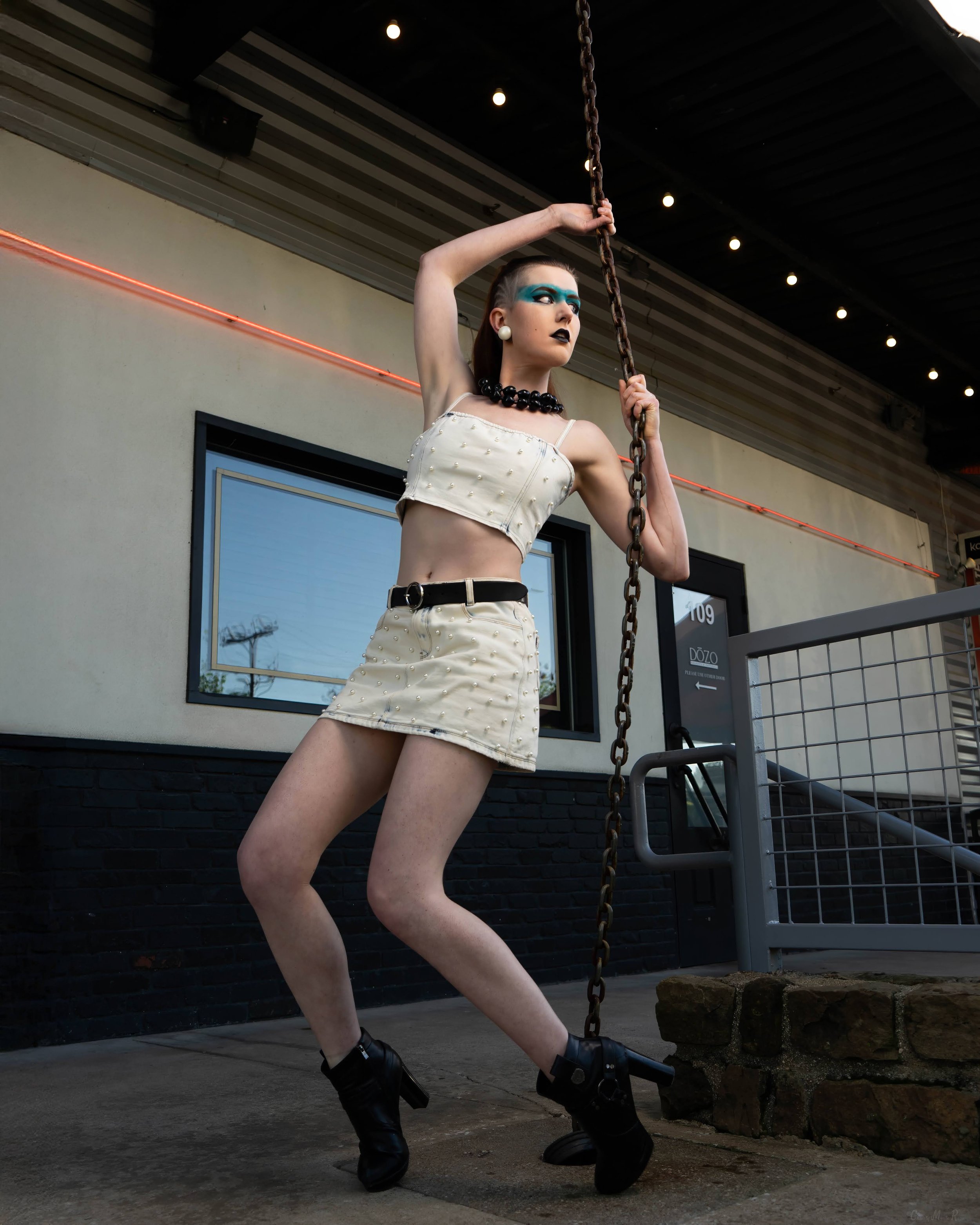 A woman with blue eyeshadow, black lipstick, and large white earrings poses outside, holding a chain with one hand and standing with her legs apart in front of a building with black brick lower wall and beige upper wall, near a window with a blue fra