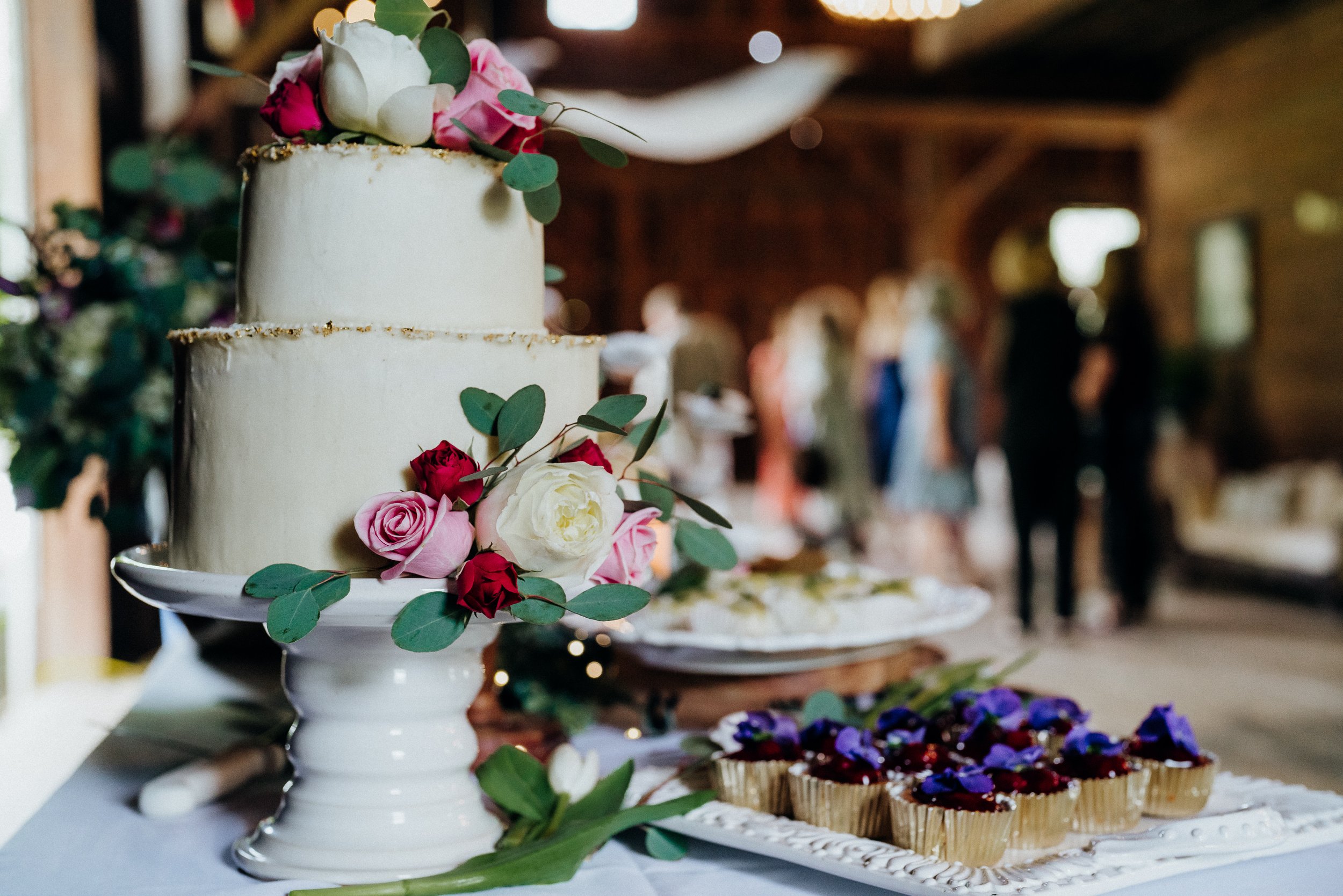 Wedding cake and other pastries on table with barn venue and guests in background