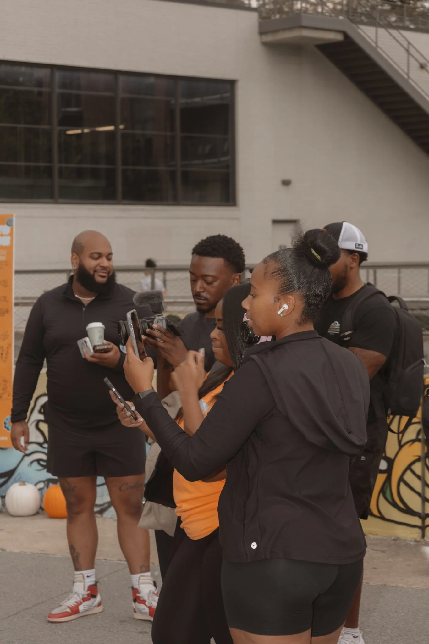 A group of people gathered outdoors, looking at their phones and a camera, with some wearing wireless earbuds and casual clothing, near a graffitied wall and pumpkins.