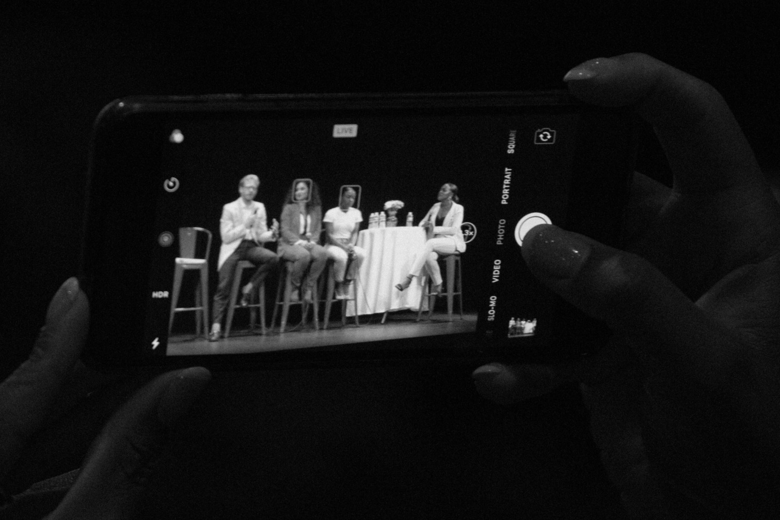 A person taking a photo of a panel discussion with four women sitting on high chairs behind a table, visible on a smartphone screen.