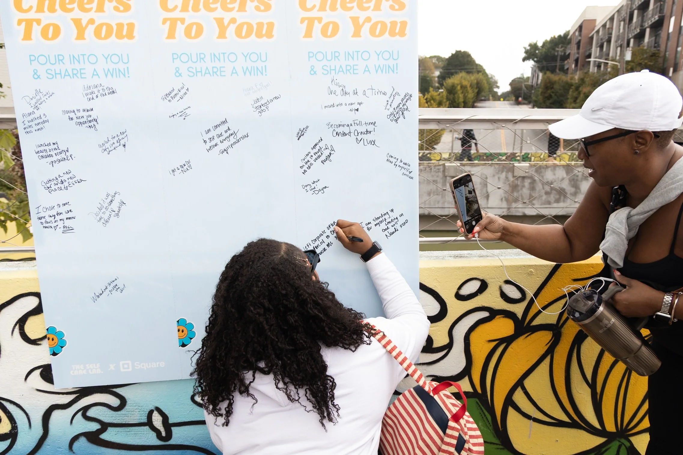 Two women writing and taking photos of a large community message board with notes and signatures, outdoors on a sunny day.