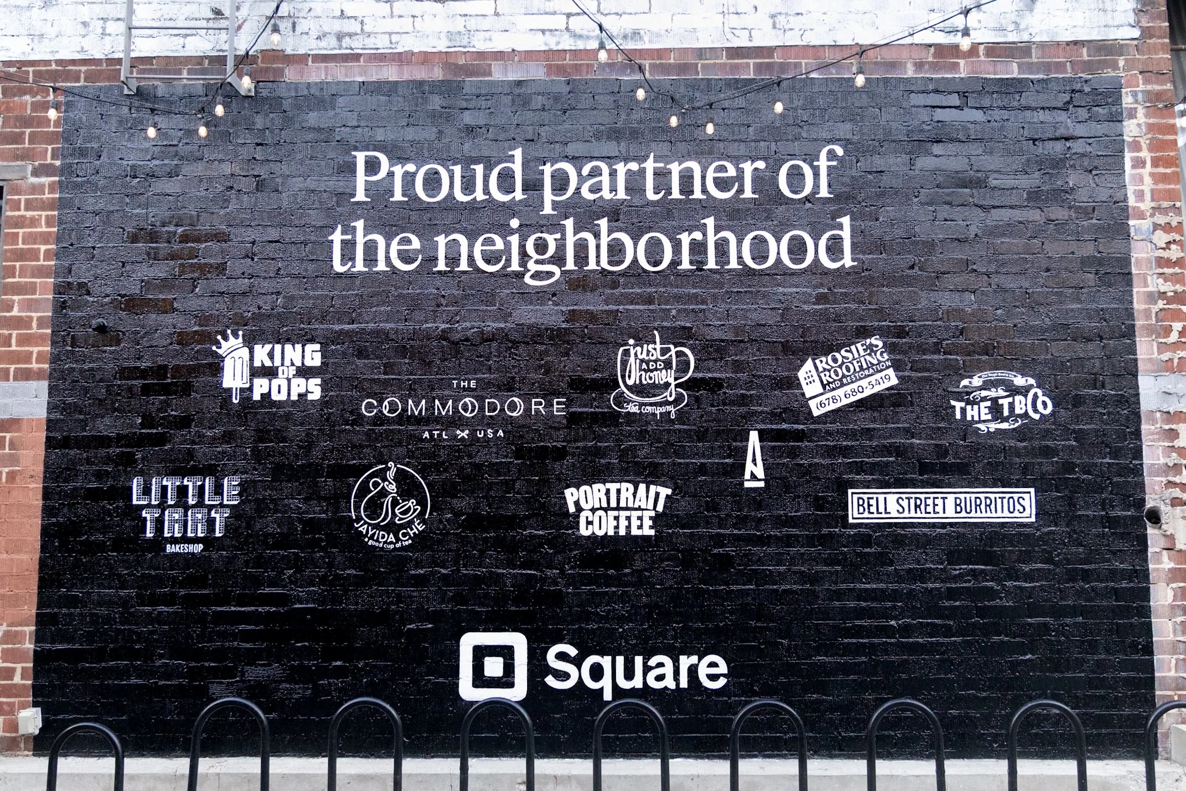 A black brick wall with white lettering that says 'Proud partner of the neighborhood' and various business logos and names, including King of Pops, The Commodore, Must Add Honey, Rosie's Roofing, The TBC, Little Tart, Jayida Che, Portrait Coffee, Bel