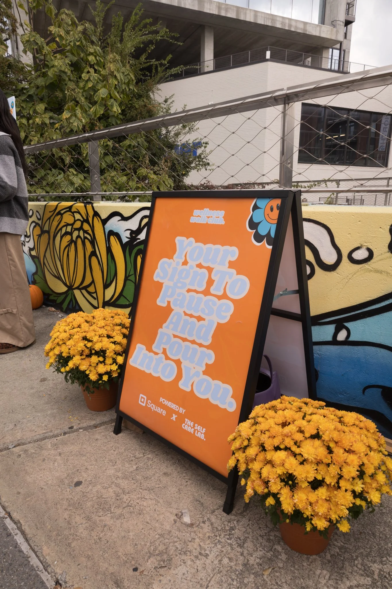An orange stand-alone sign with white text that reads 'Your Sign To Pause And Pour Into You.' Two yellow potted chrysanthemums are placed on either side of the sign, on a sidewalk next to a colorful mural wall. A person wearing beige pants and a dark