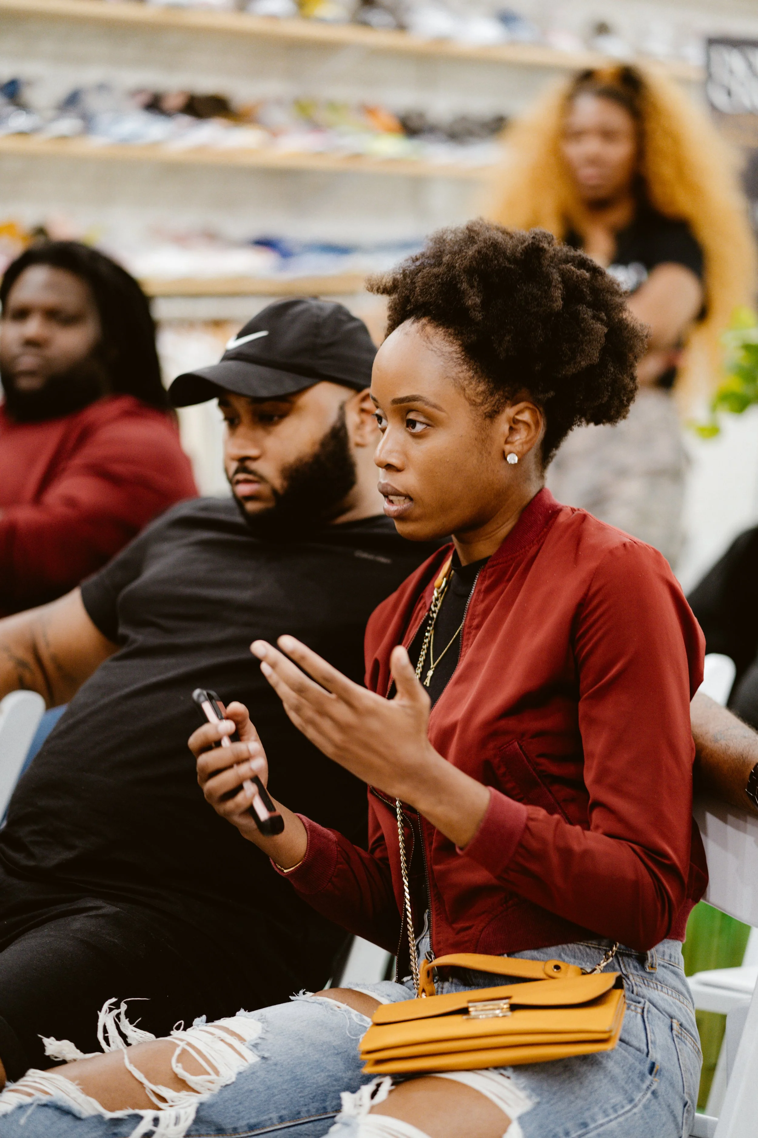 A woman with short, curly hair wearing a red jacket, ripped jeans, and holding a phone sits participating in a discussion in a group setting.