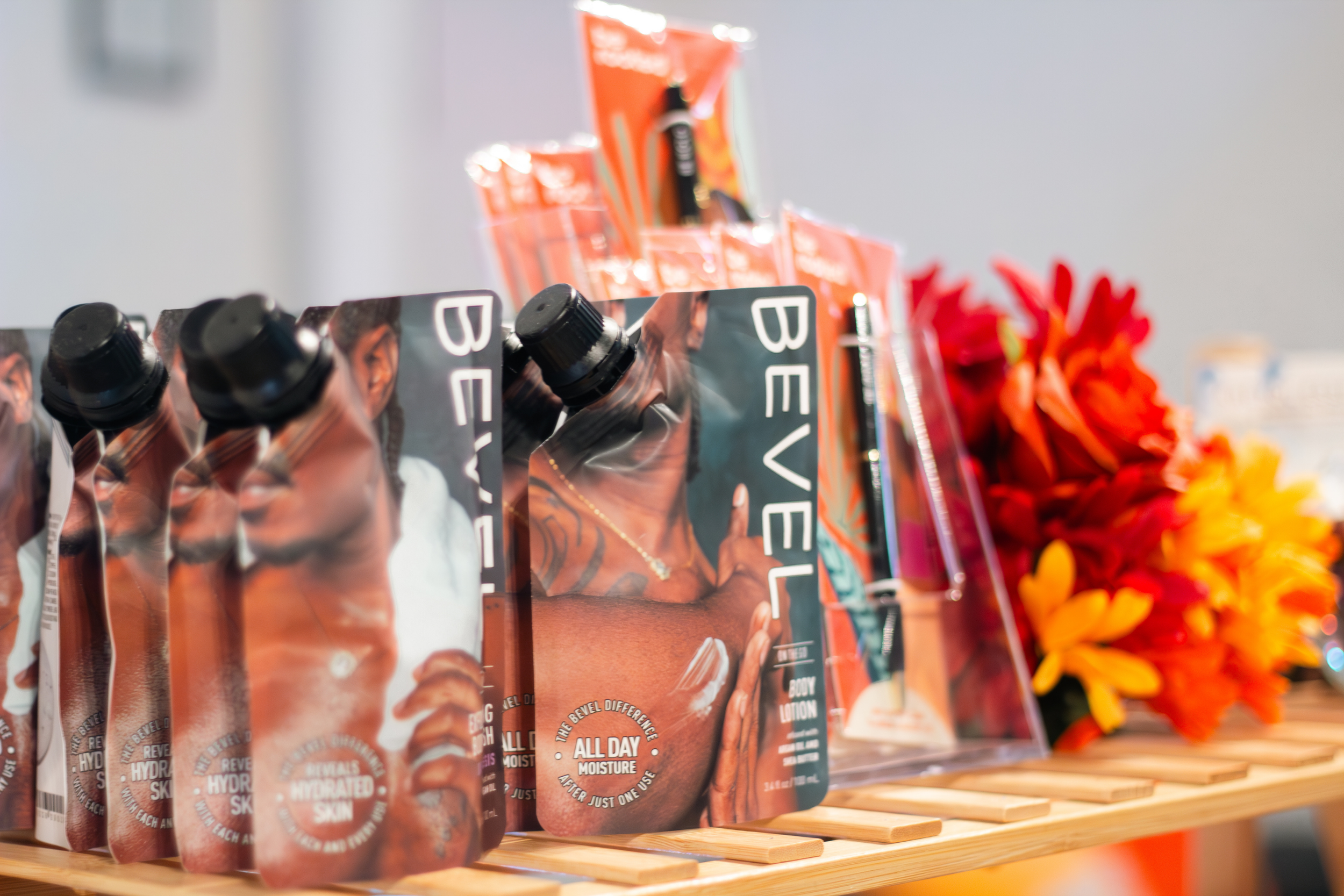 Personal care products including BEVEL body lotion in squeeze pouches, displayed on a wooden surface with orange and red flowers in the background.