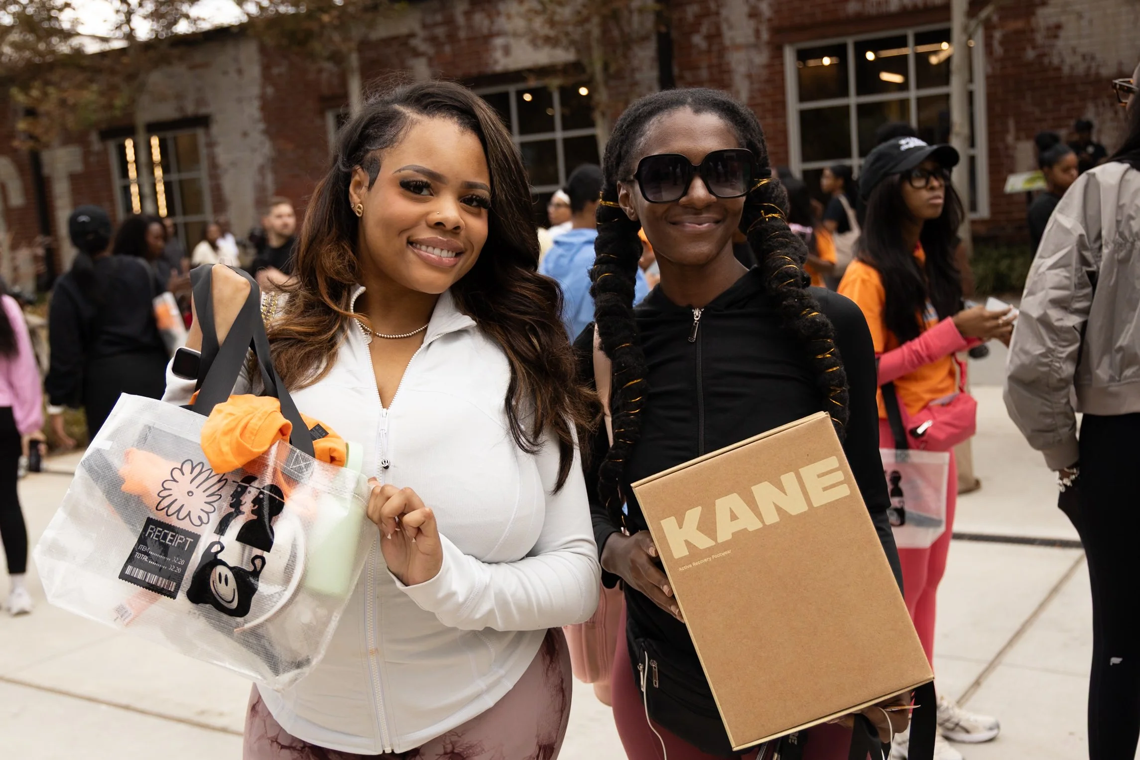 Two women standing outdoors at an event, smiling at the camera. One woman wears a white zip-up jacket and holding a clear tote bag, the other wears a black hoodie and large sunglasses, carrying a KANE shoe box.