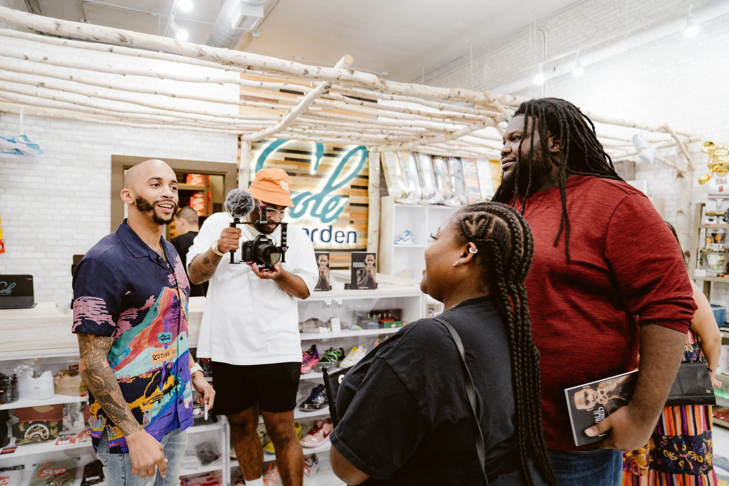 Four people in a retail store, two men and two women, engaging in a conversation. One man is holding a camera with a stabilizer, and another man is holding a magazine. The store has a sign that reads 'Eagle' and shelves with various shoes and merchandise.