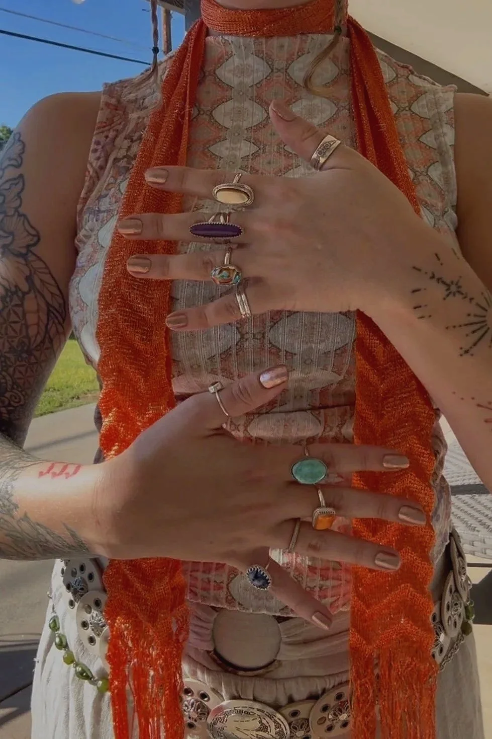 Close-up of a person's hands with numerous rings, showing tattoos on the arms, wearing patterned clothing and an orange scarf, with a clear sky in the background.