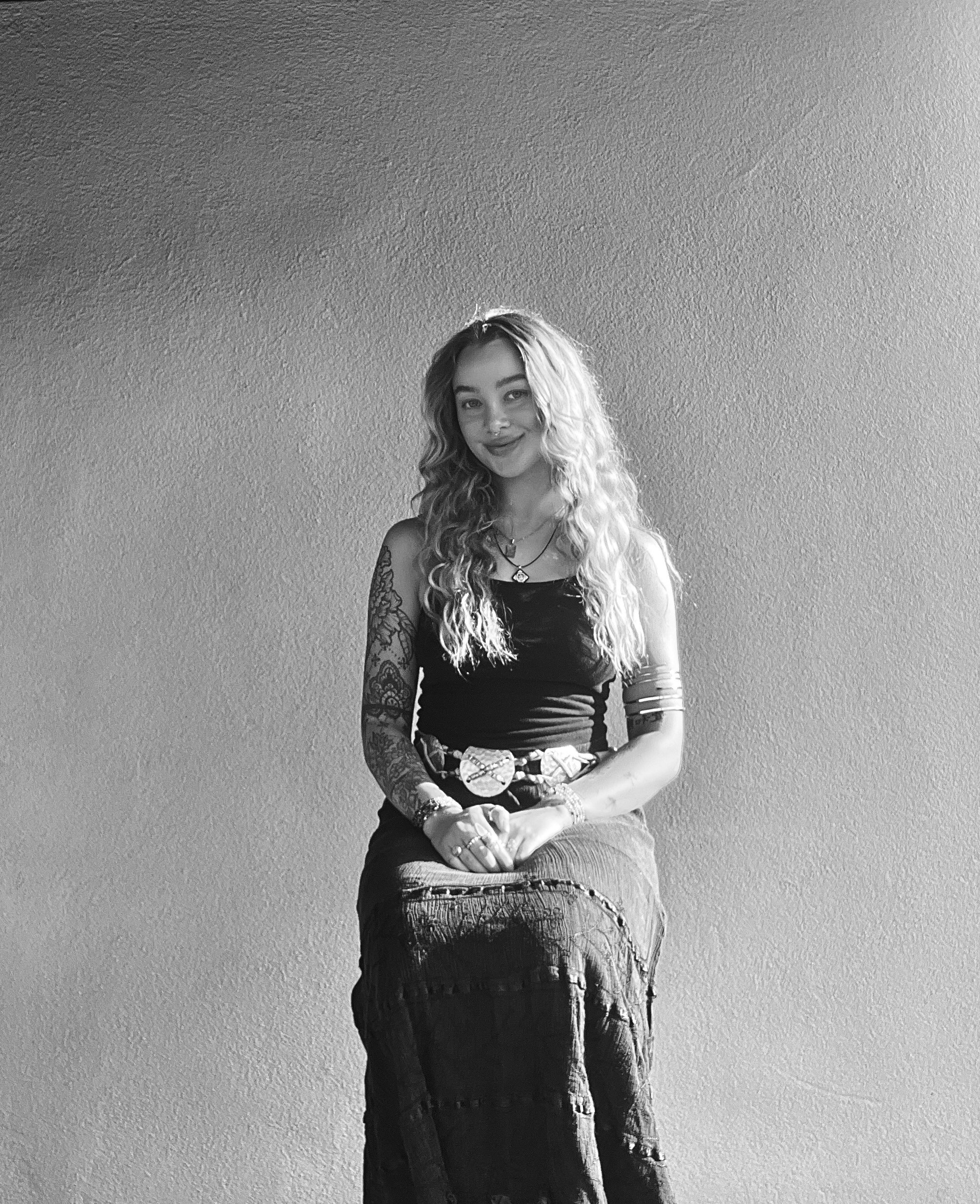 A young woman with long curly hair, wearing a sleeveless top and long skirt, sitting and smiling in front of a plain wall.