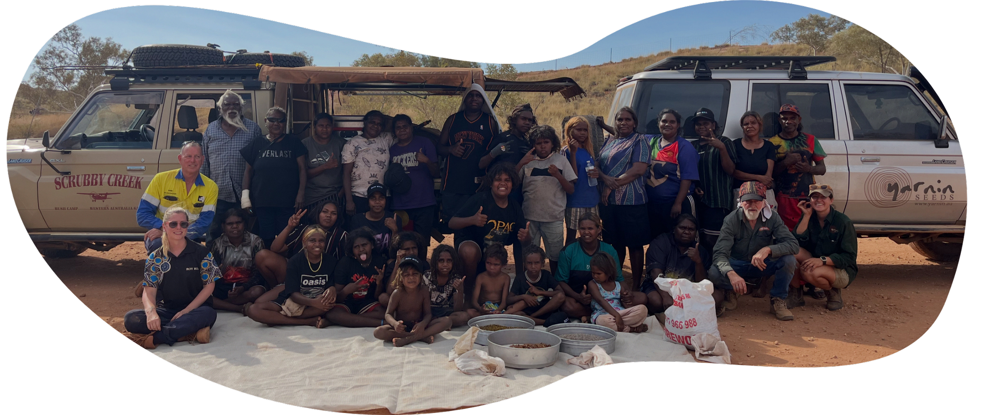A group of Yarnin Seeds workshop participants posing in front of two off-road vehicles, with a white tarp and collections of seed on the ground, in the Pilbara.