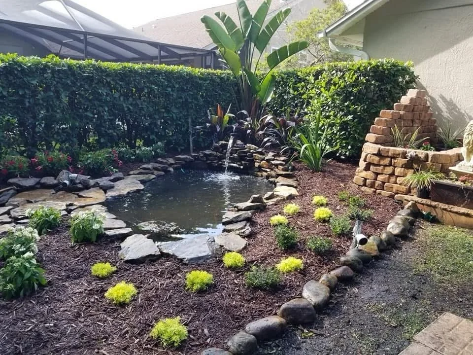 Backyard garden with a small pond surrounded by rocks, lush green plants, and a hedge. There is a small waterfall feature and a decorative brick structure on the right.