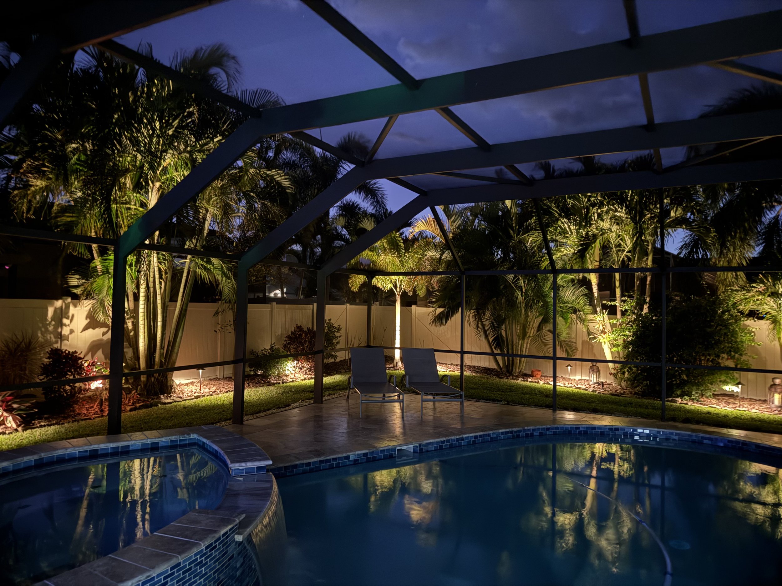 Night view of a house with exterior lights illuminating the front entrance and landscaping, including palm trees and pathway lighting.